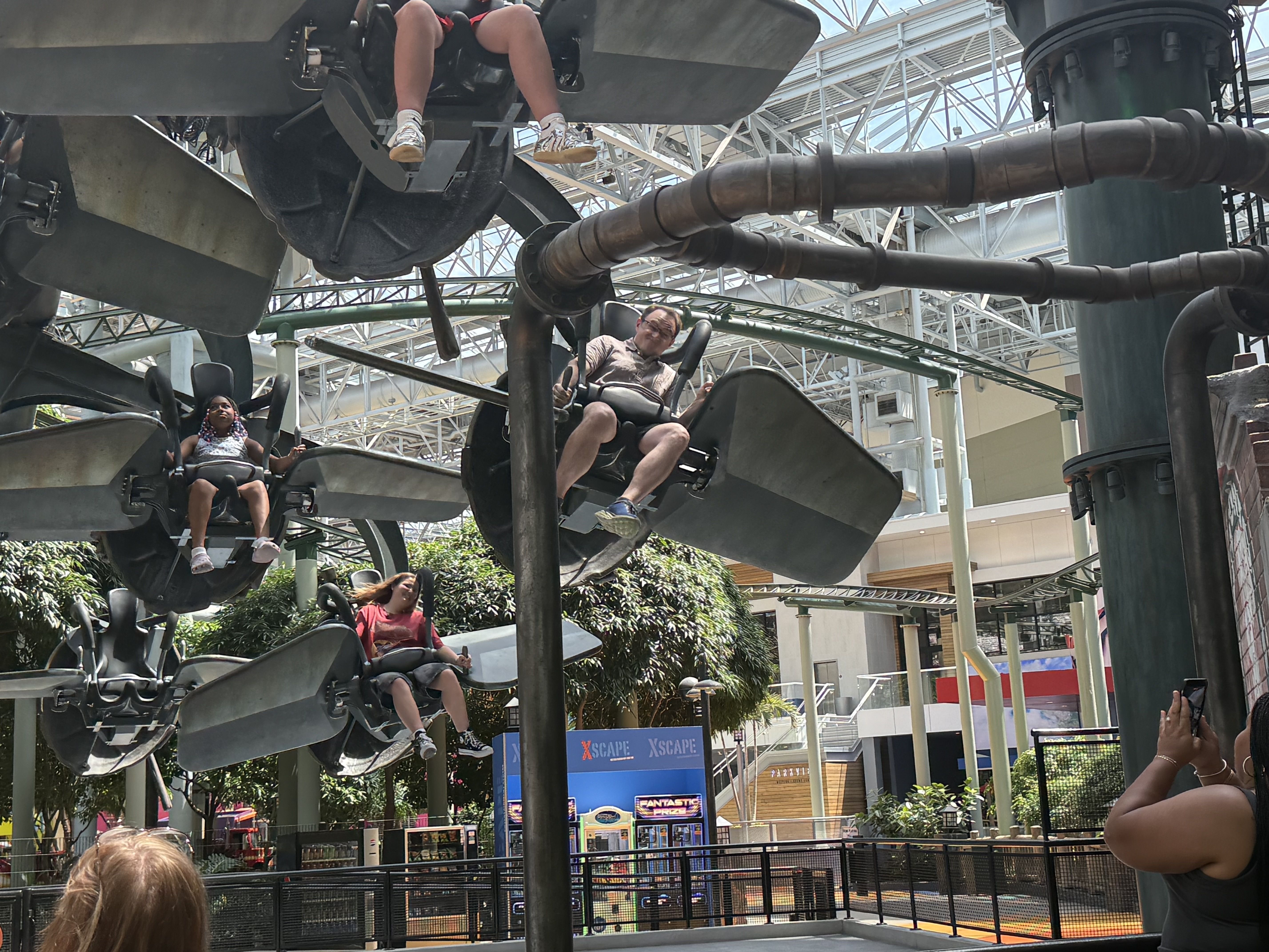 People ride a suspended roller coaster in a bright indoor amusement area; two riders visibleโman with glasses and a womanโseated in open seats, with metal track and glass roof overhead.