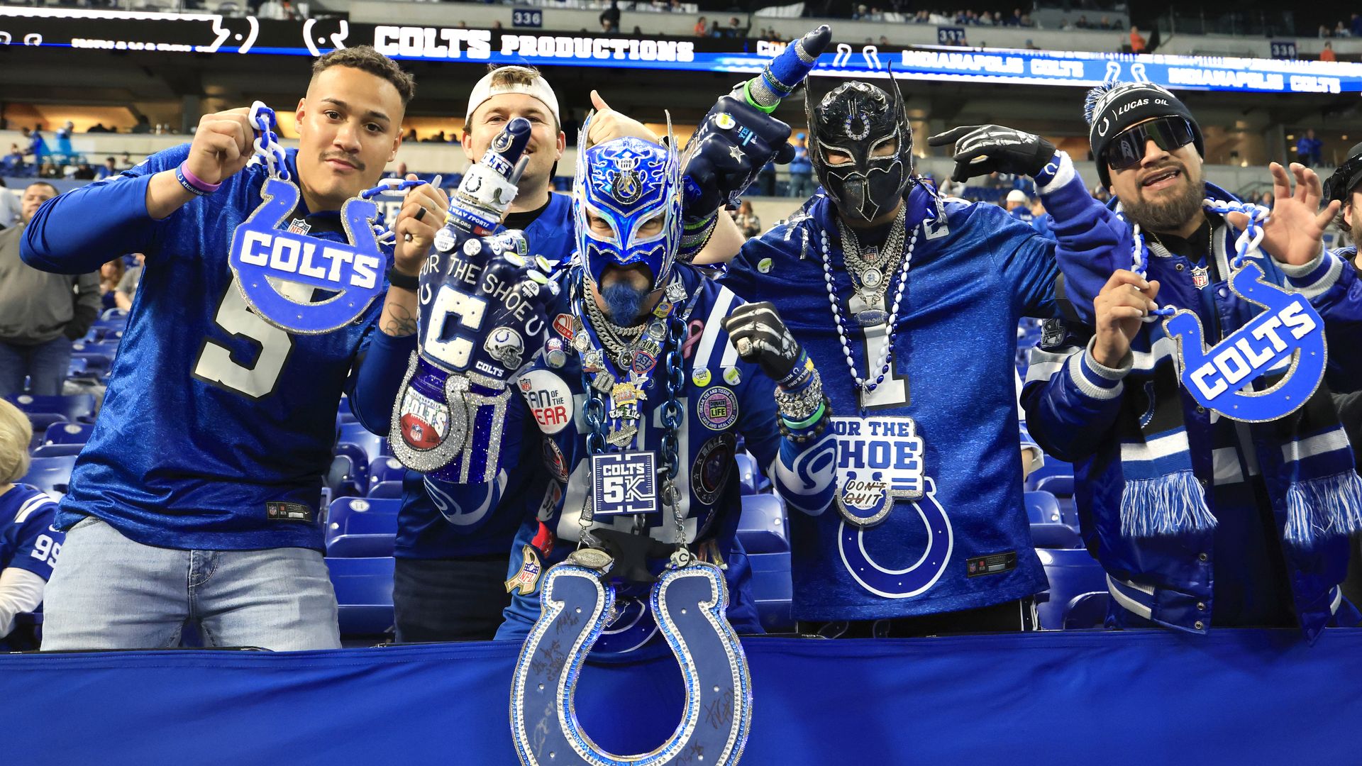 Five enthusiastic fans dressed in blue Indianapolis Colts gear, some wearing masks and large Colts holiday chains, posing inside a stadium with Colts signage in the background.