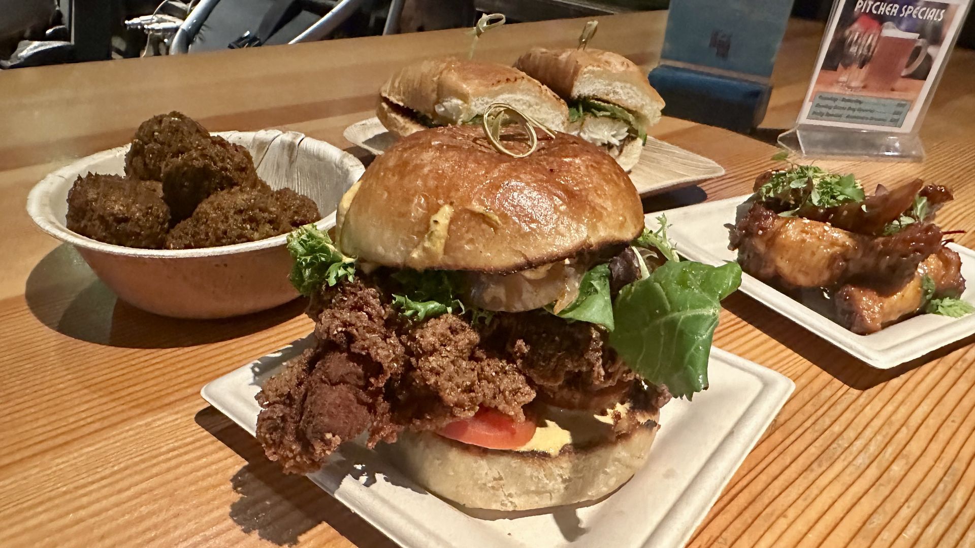 A large fried chicken sandwich in the foreground, plus chickpea fritters to the left a catfish po' mi in back, and berbere whisky glazed chicken wings at right, sitting on a wooden table.
