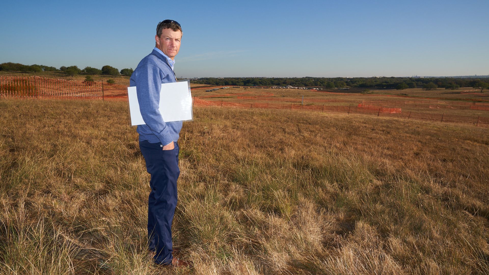 a person in blue sweater and pants holding whiteboards, standing in dry grassy field with orange construction fencing and trees in the distance under clear blue sky.