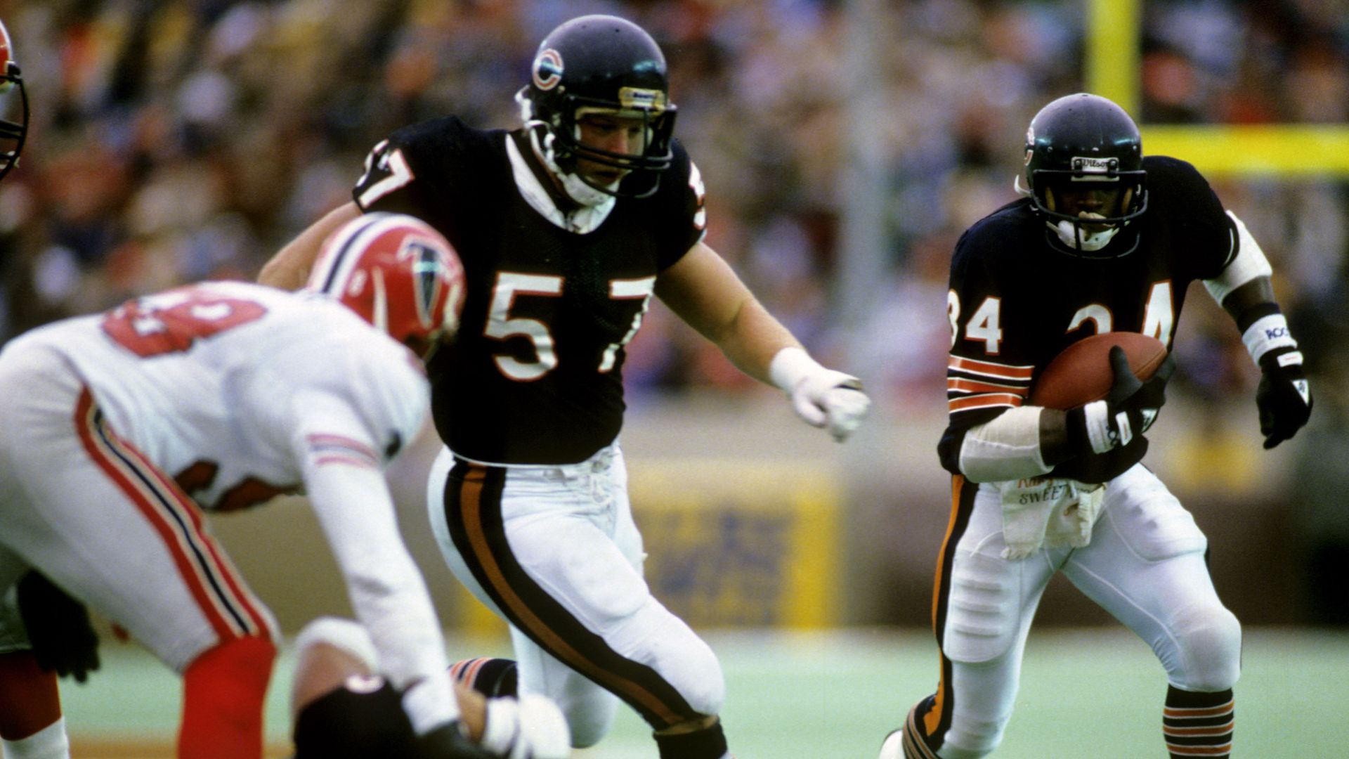 The back of an Atlanta Falcons player, bent over, next to Chicago Bears' Tom Thayer in Bears uniform running alongside Walter Payton as Payton runs the ball down the field.