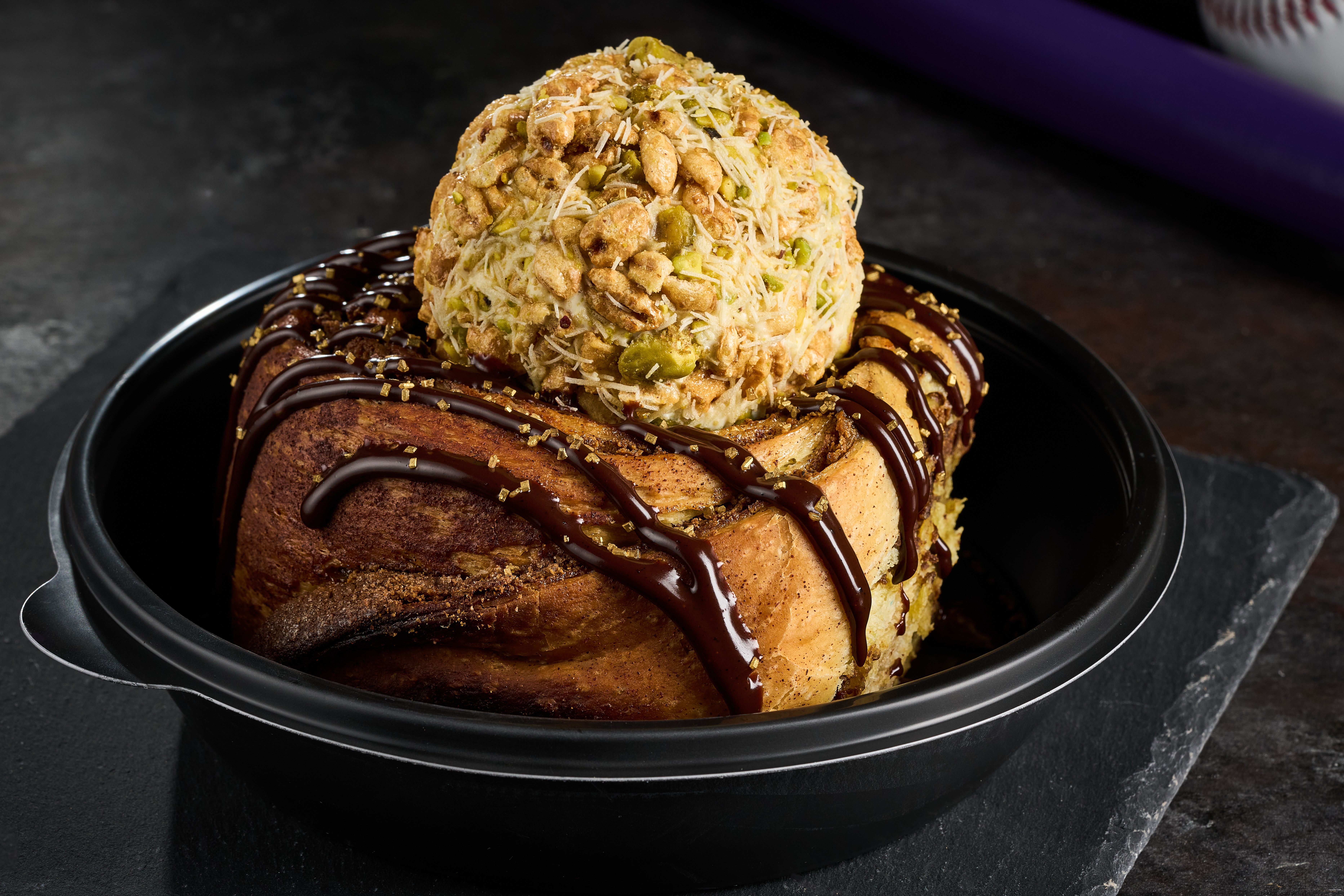 A large cinnamon-roll loaf drizzled with dark chocolate and gold sugar, topped by a coconut-pistachio ball, all in a black baking dish.