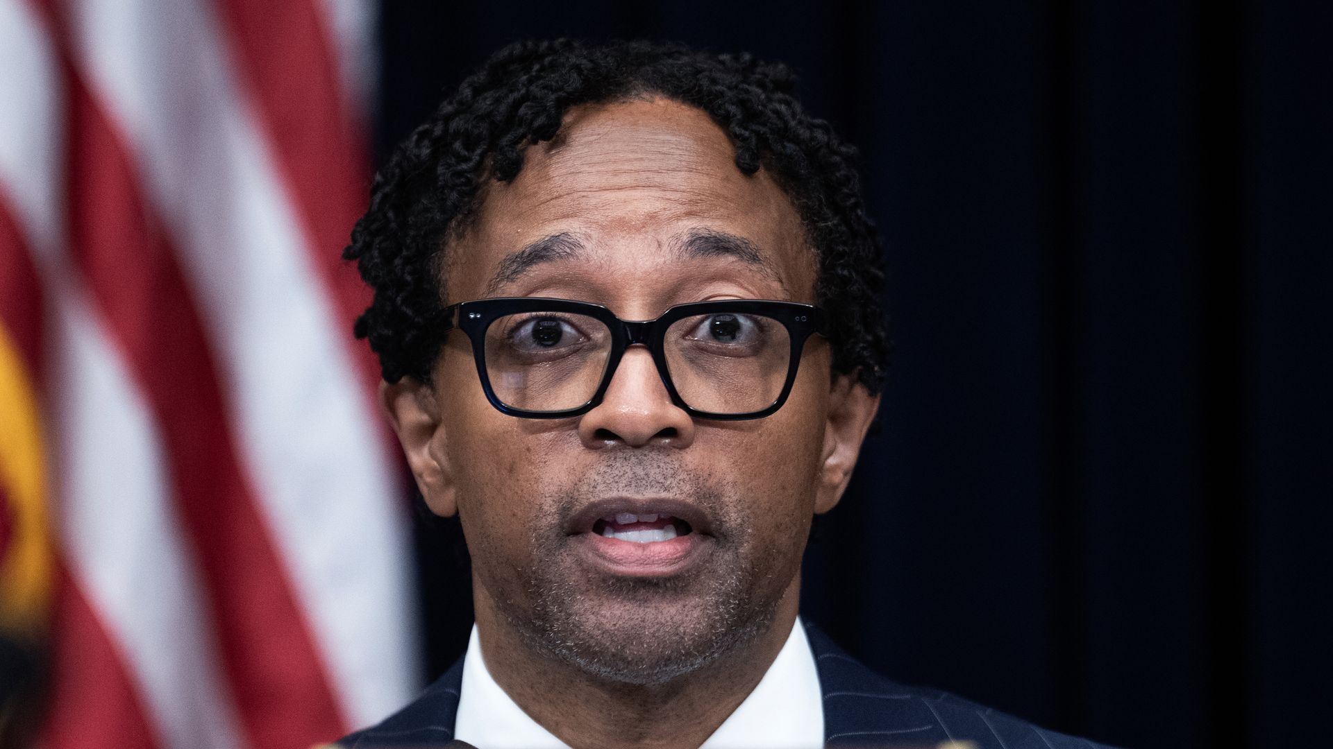 Close-up of a man with curly hair and black-framed glasses, speaking at a podium in a dark pinstripe suit and white shirt; a blurred red-and-white flag is behind him.