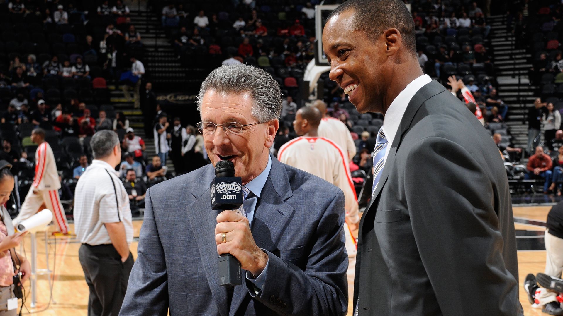 Two men smile at something off camera while standing on the Spurs basketball court. 