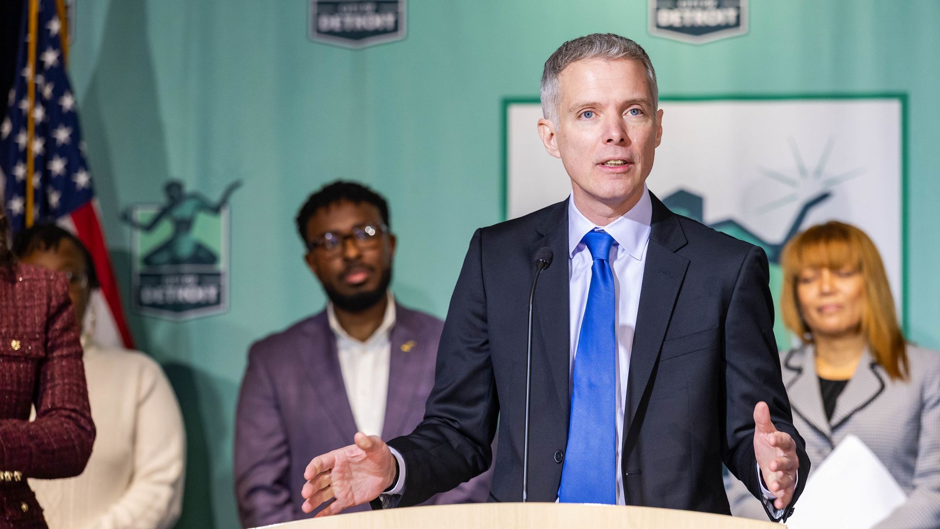 Man in black suit and blue tie speaking at podium with microphone, three people behind him.