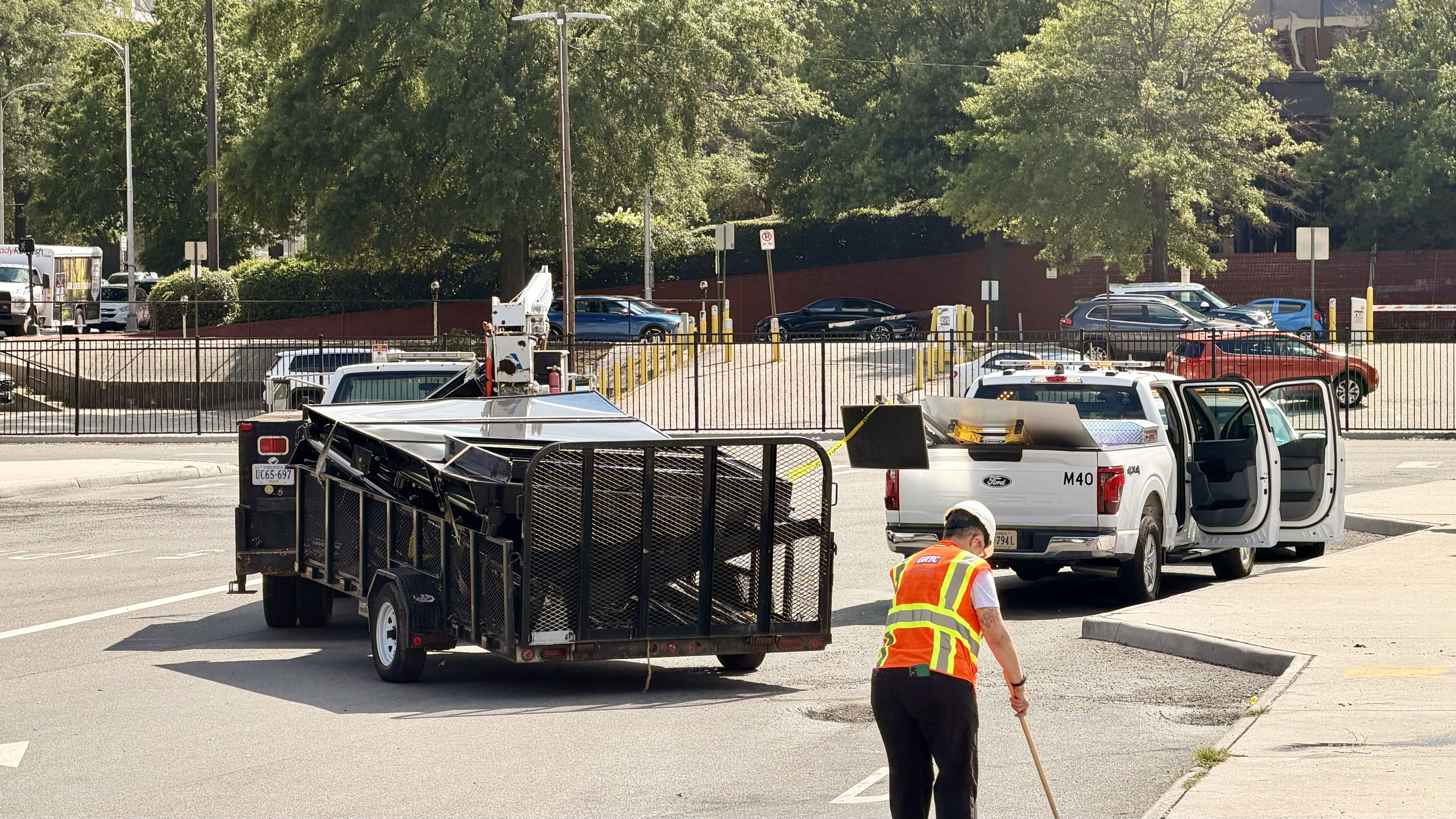Sunlit parking lot with a white Ford pickup and a black trailer in the foreground; a worker in a neon orange safety vest stands near the truck while cars and green trees line the background.