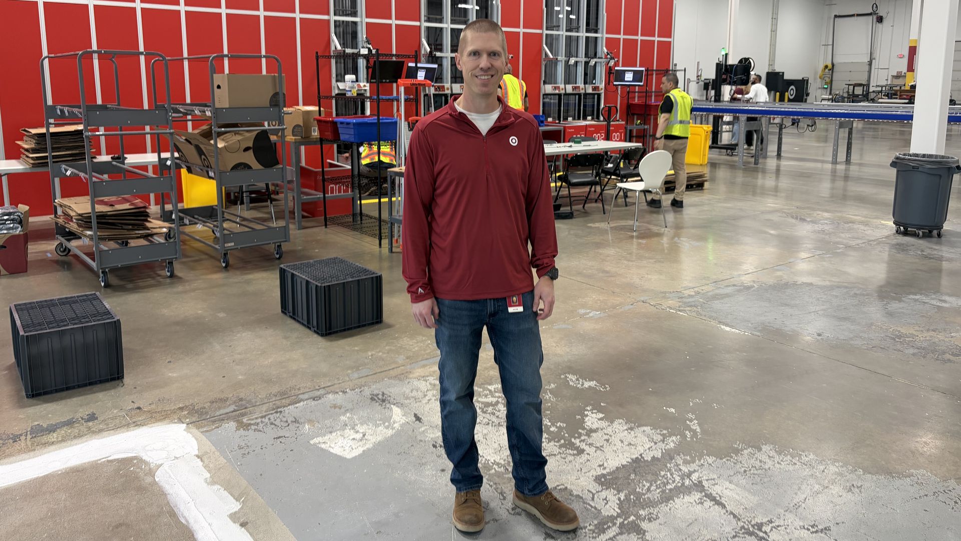 Jake Krueger in a maroon shirt stands in a large industrial workspace with red wall panels, metal racks and carts. In the background, workers in safety vests operate equipment.