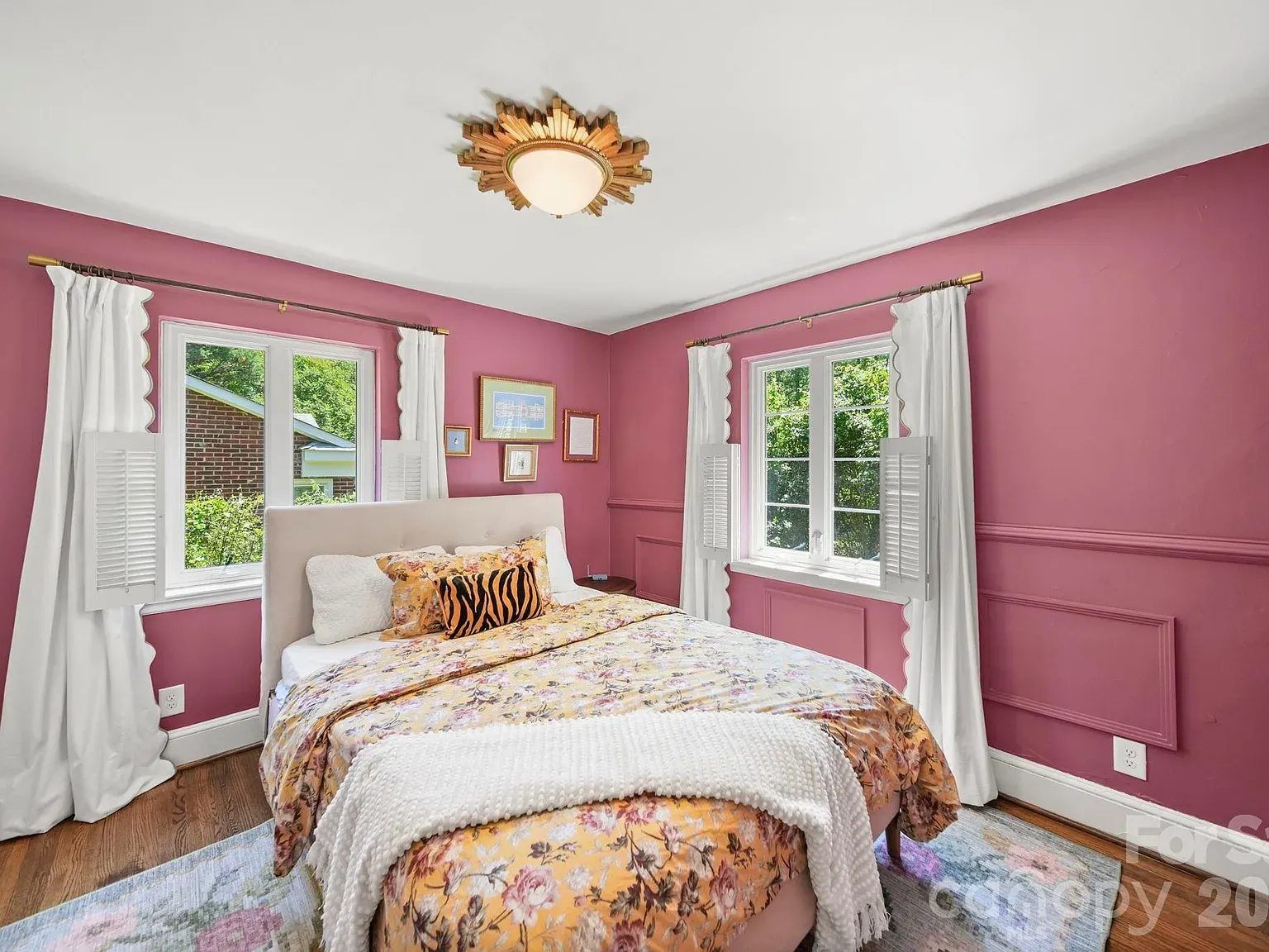 Bright pink bedroom with white curtains, a light upholstered bed in front of two windows with shutters, floral quilt, zebra pillow, wall art, and a sunburst gold ceiling light.