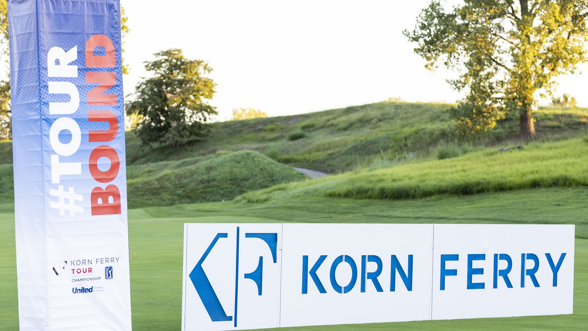 A tall vertical blue banner on the left with large white text and red 'TOUR' lettering; white boards in the foreground read 'KORN FERRY' in blue, set on a green grassy hill with trees and a bright sky.
