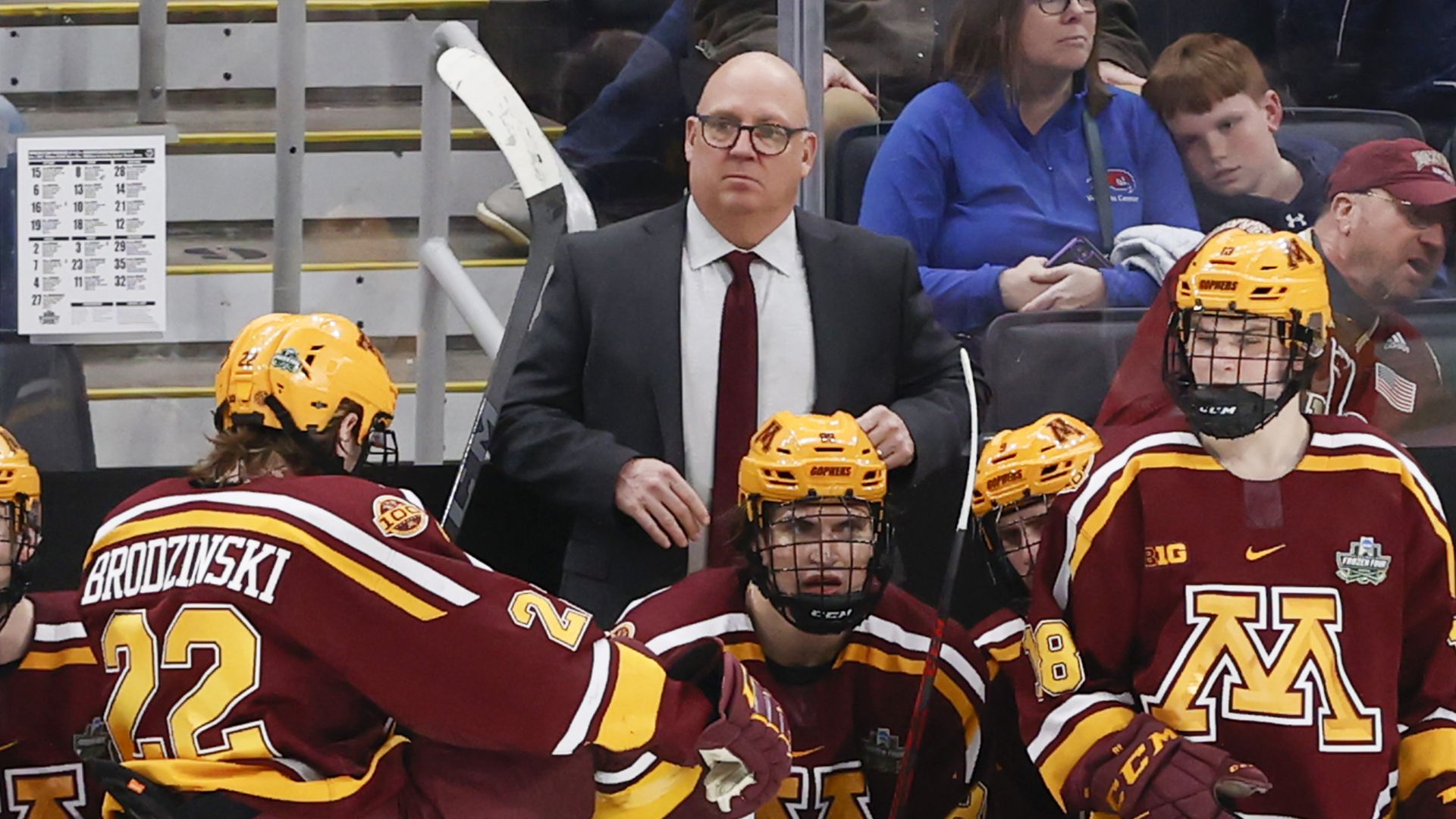 Hockey players in maroon and gold uniforms with yellow helmets sit on the bench; a bald coach in a suit stands behind them. Glass divider and spectators visible in the background. 