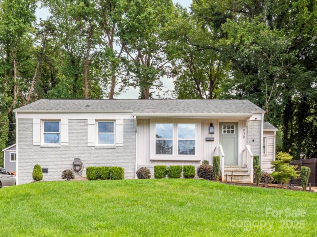 Single-story gray brick house with green front door, white window shutters, small shrubs in front, and large green lawn surrounded by tall trees, listed for sale in 2025.