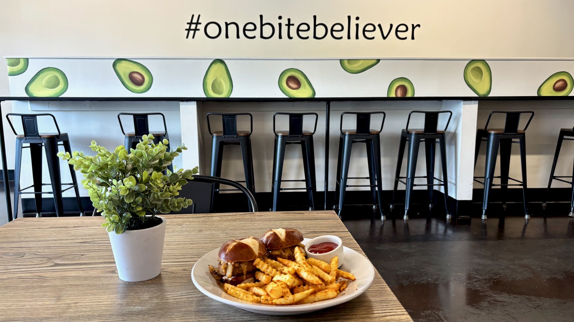 Cafe interior with avocado graphics and the hashtag #onebitebeliever. Black metal bar stools line a long counter; a potted plant sits on a wooden table; plate with sandwich and fries in foreground.
