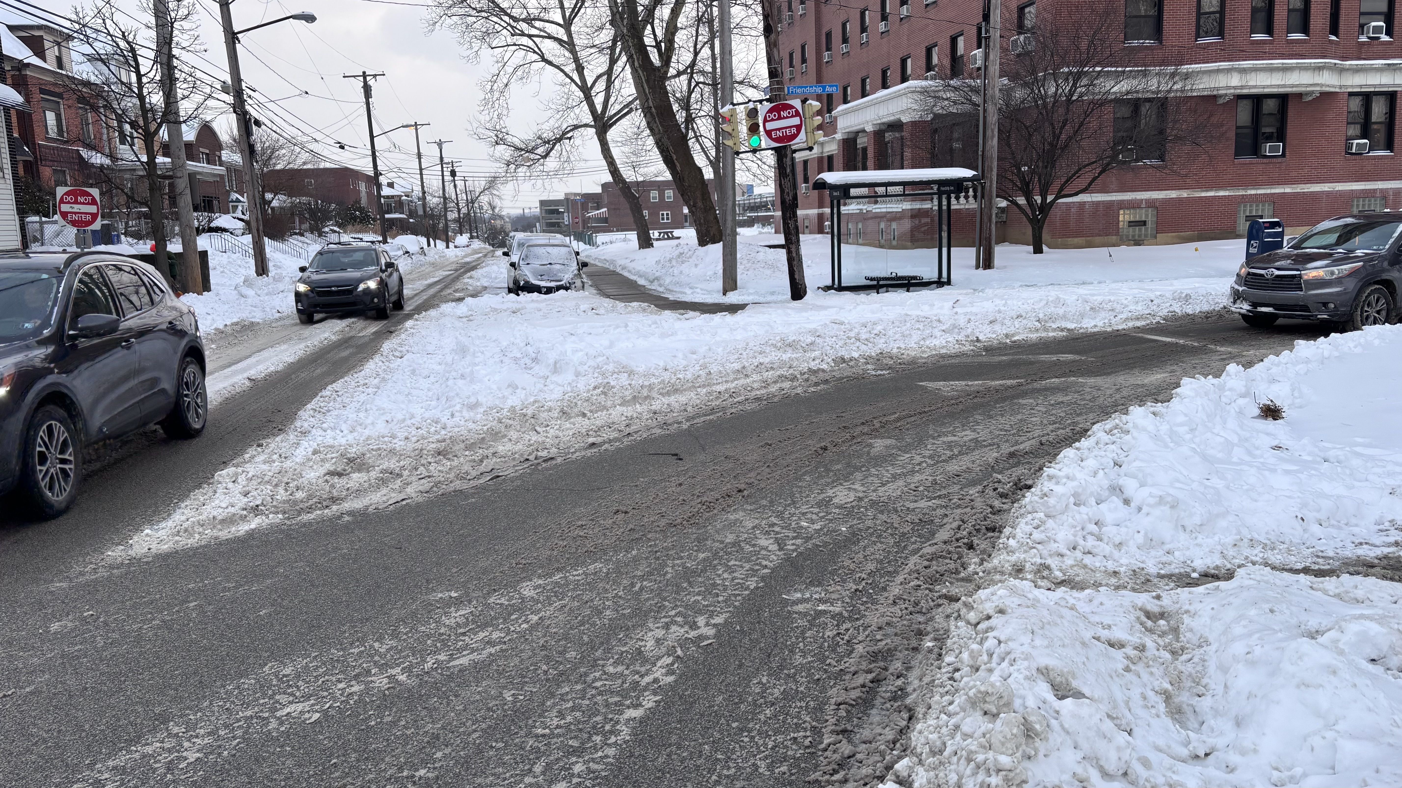Snow-covered street intersection with cars, slushy roads, a bus stop shelter, and "Do Not Enter" signs on lampposts in a cloudy winter day urban neighborhood.