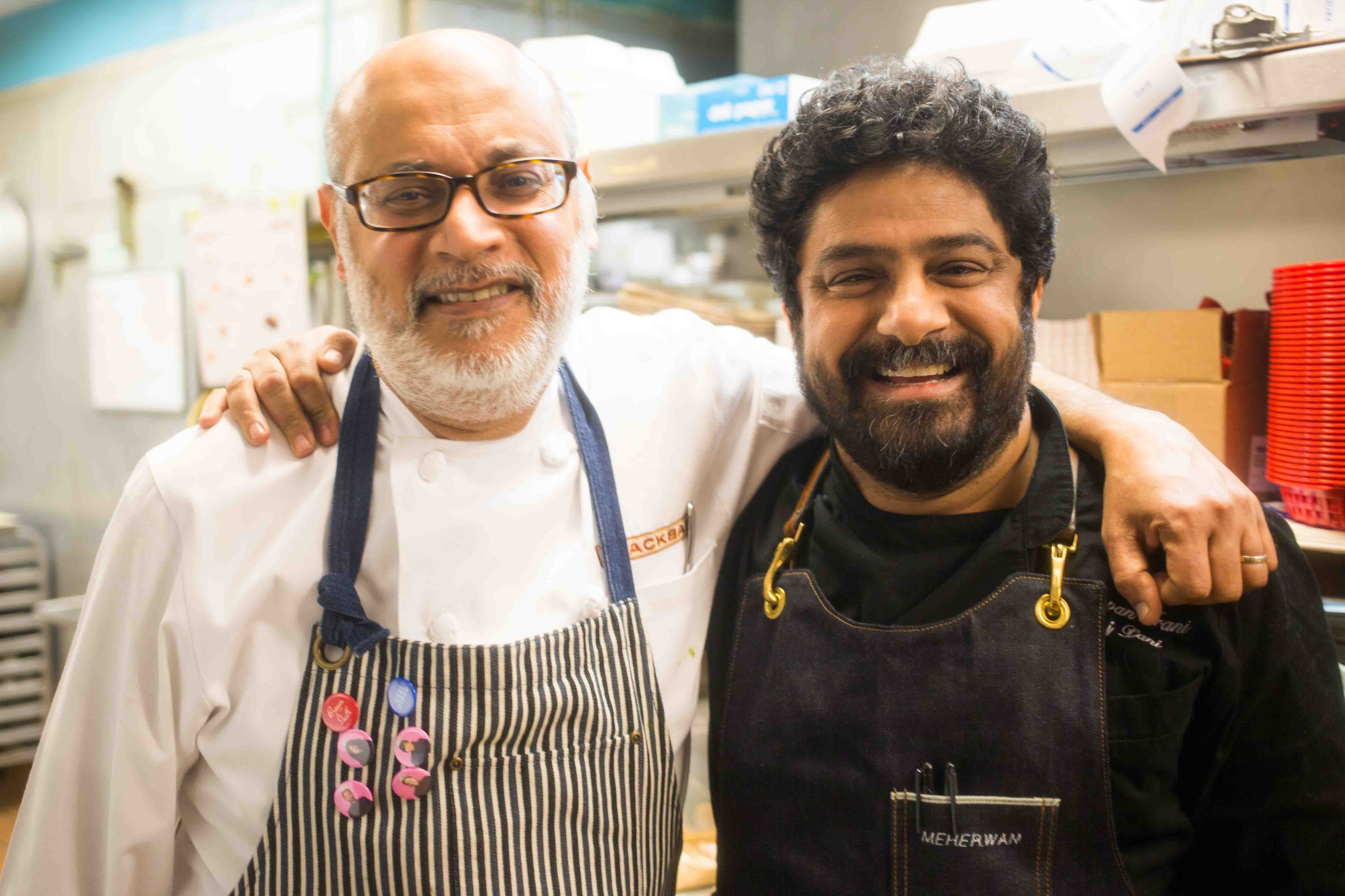 Two male chefs smiling with arms around each other in a kitchen, one wearing glasses, white coat, striped apron with pink badges, the other in black coat with dark apron labeled "Meherwan".