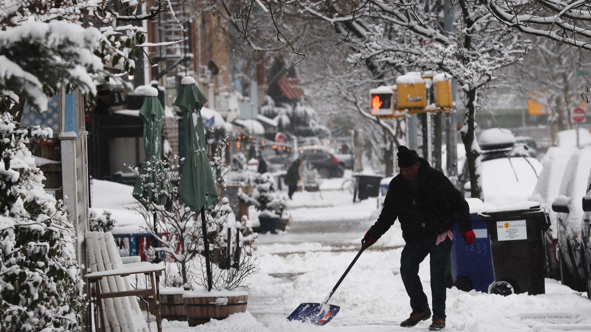 The silhouette of a man shoveling snow from a path in Brooklyn, New York City, with snow-covered trees and parked cars to his left and trees and buildings to his right.