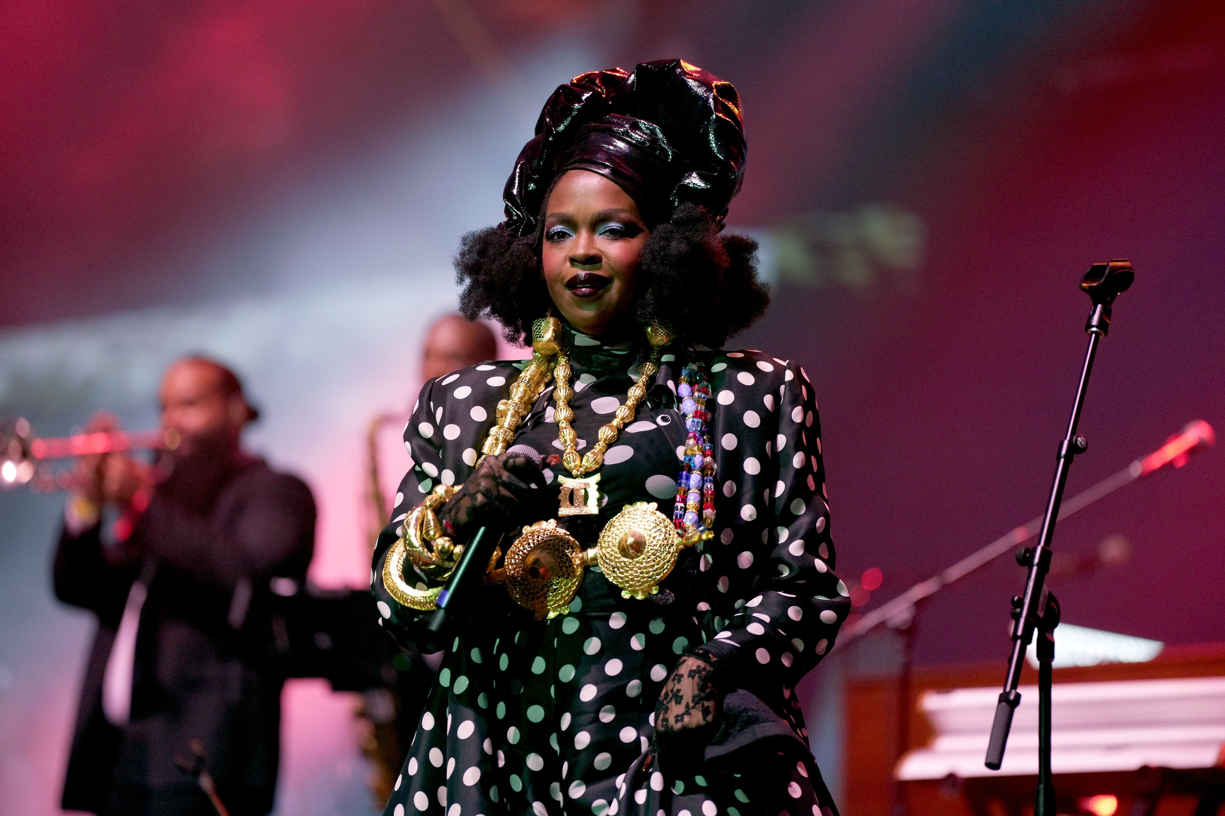 Singer on stage wearing a black and white polka dot outfit, large gold necklaces, black lace gloves, and a shiny black headwrap, holding a microphone with a band playing in the background.
