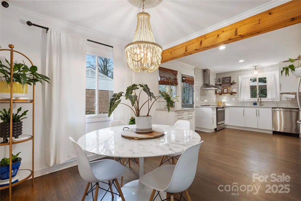 Bright kitchen and dining area with white cabinets, marble round table, white chairs, wooden floor, a plant centerpiece, gold chandelier, and large wooden beam ceiling. For Sale sign.