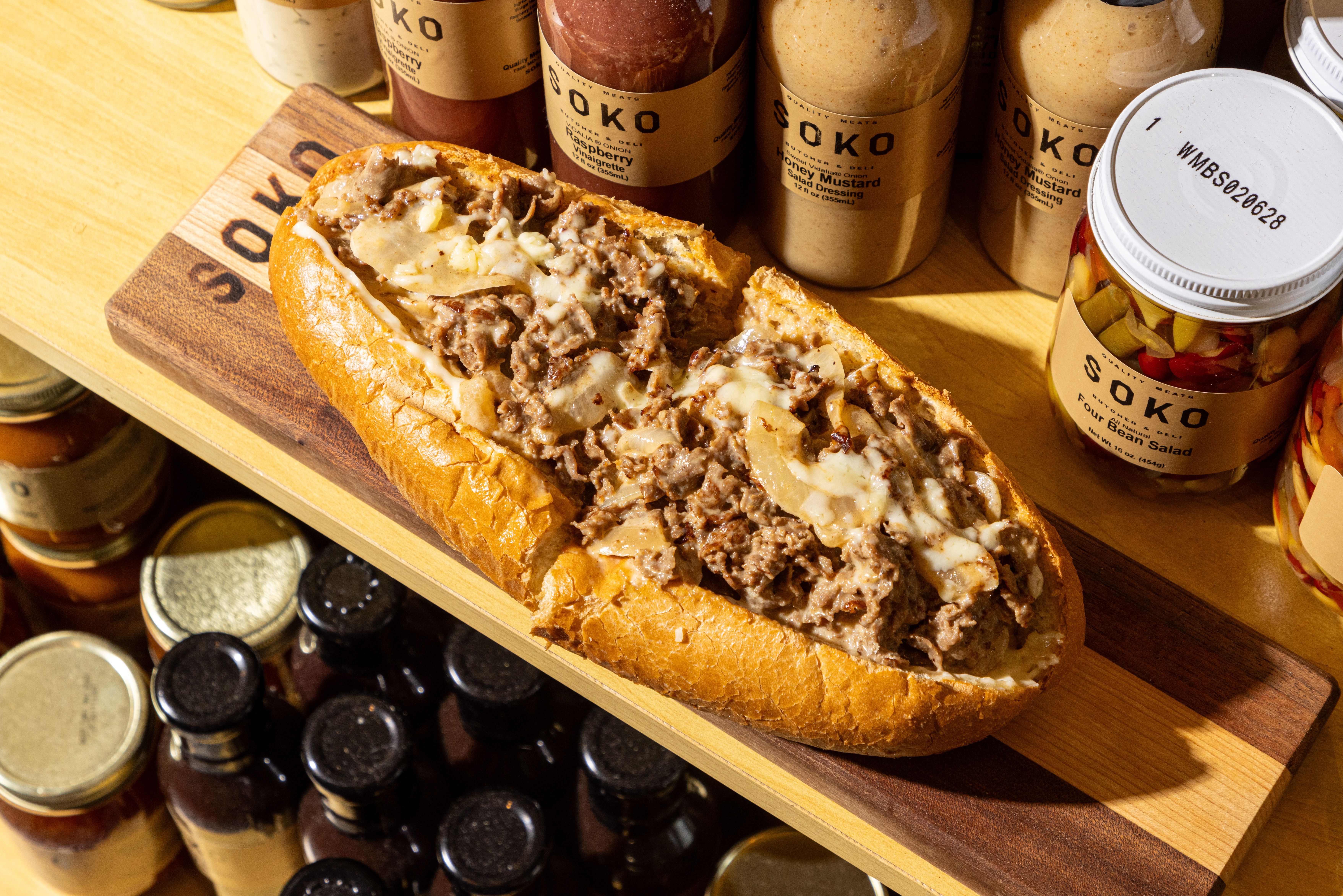 A beef sandwich with melted cheese on a crusty baguette rests on a wooden cutting board in a pantry, surrounded by sauce bottles and pickled jars.