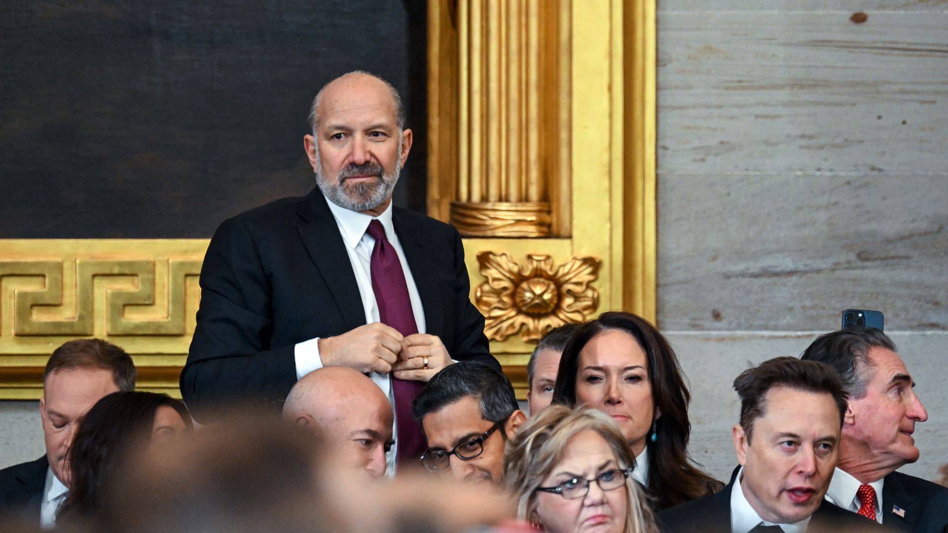 A person in a suit and tie stands amid a seated crowd