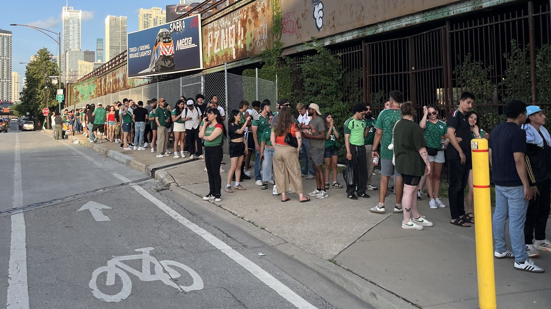 A long line of people on a sidewalk next to green fence next to a bike lane with a bike and arrow painted in white