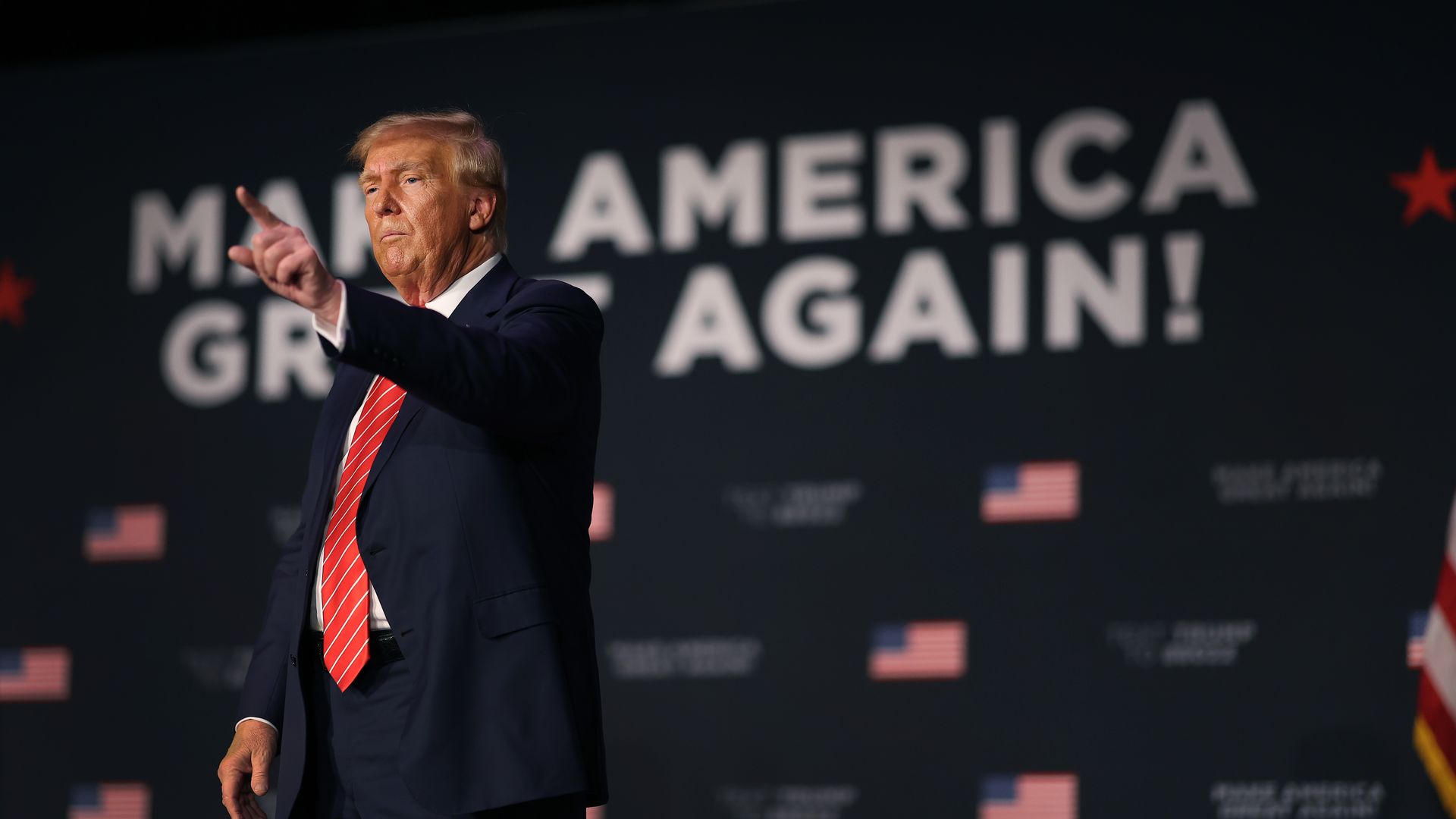 Trump at a rally with a hand outstretched pointing off camera. A sign in the background says "make America great again" in all caps with American flags.