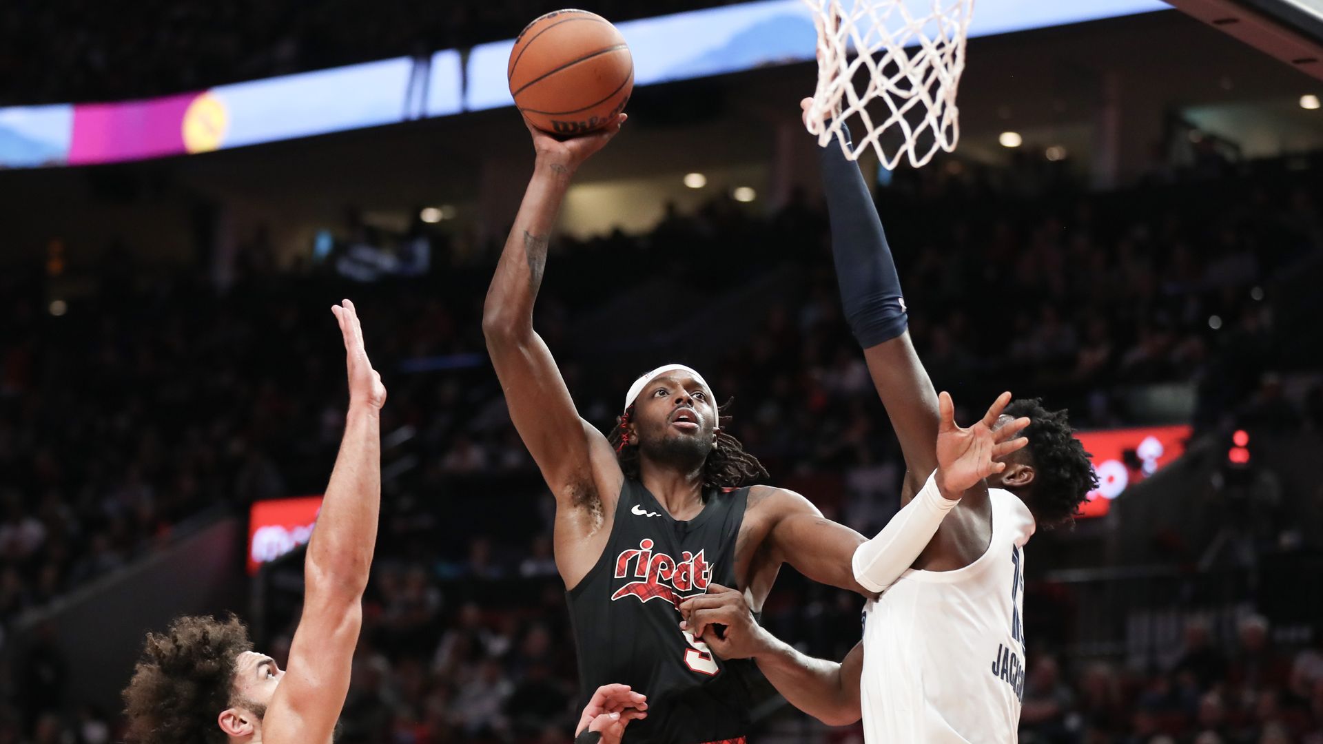 Jerami Grant #9 of the Portland Trail Blazers  holds a basketball with his fully extended right arm, going up for a shot in between David Roddy #21 and Jaren Jackson Jr. #13 of the Memphis Grizzlies, who are jumping with one arm outstretched on either side of Grant. 