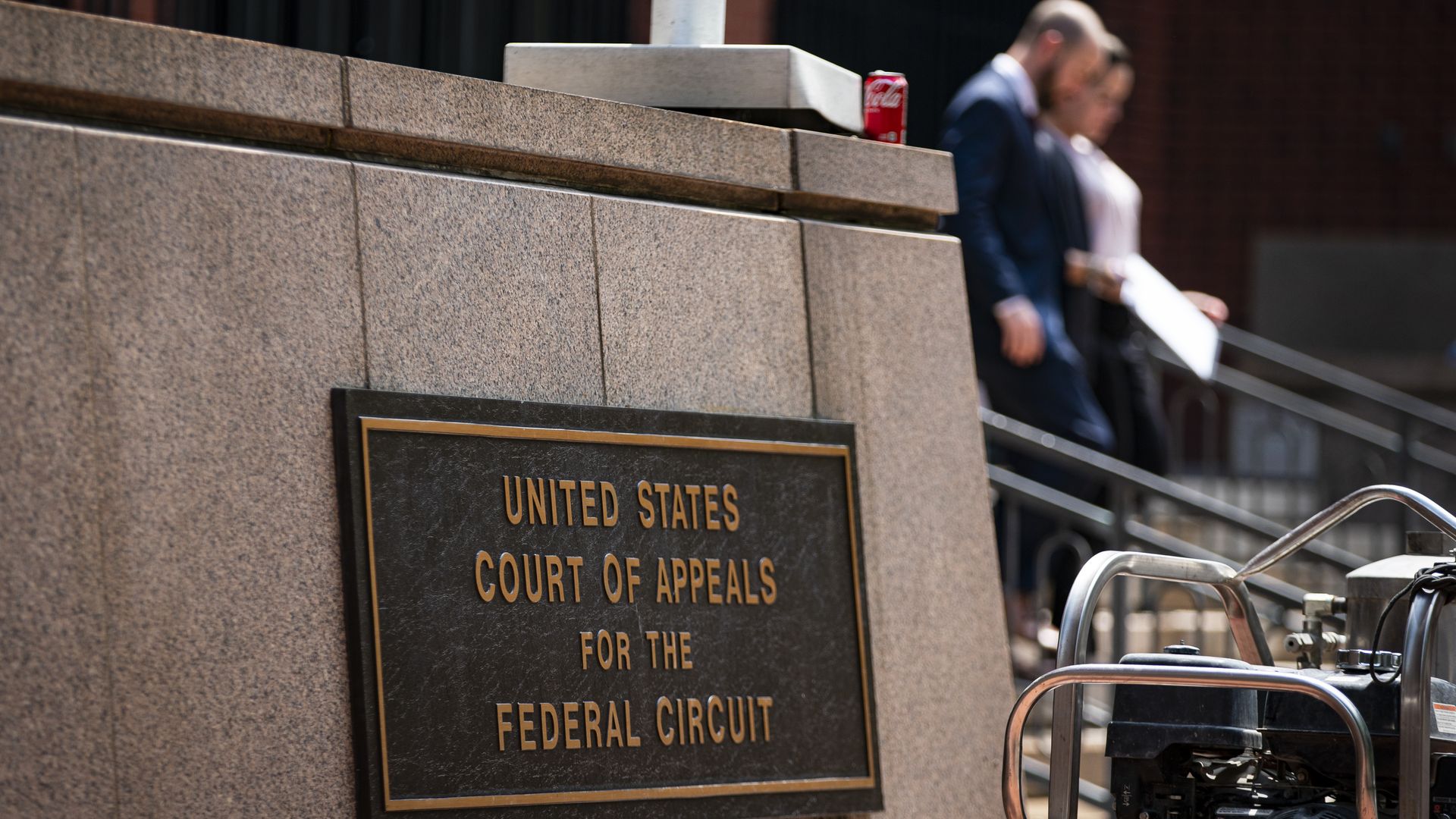 Plaque on stone wall reading "United States Court of Appeals for the Federal Circuit," with two blurred people walking down stairs and a red Coca-Cola can on top of the wall.