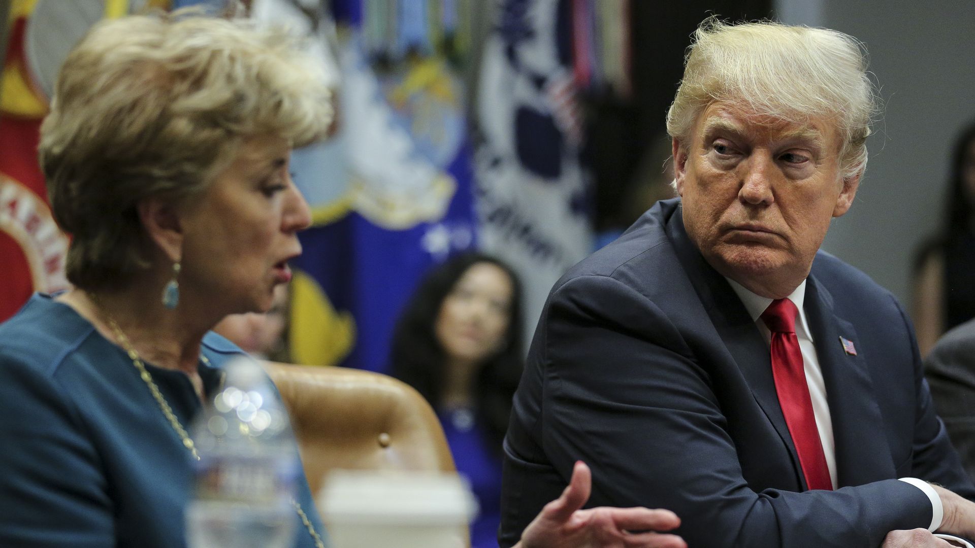 In this image, Linda McMahon talks at a meeting and Trump sits next to her, listening. 