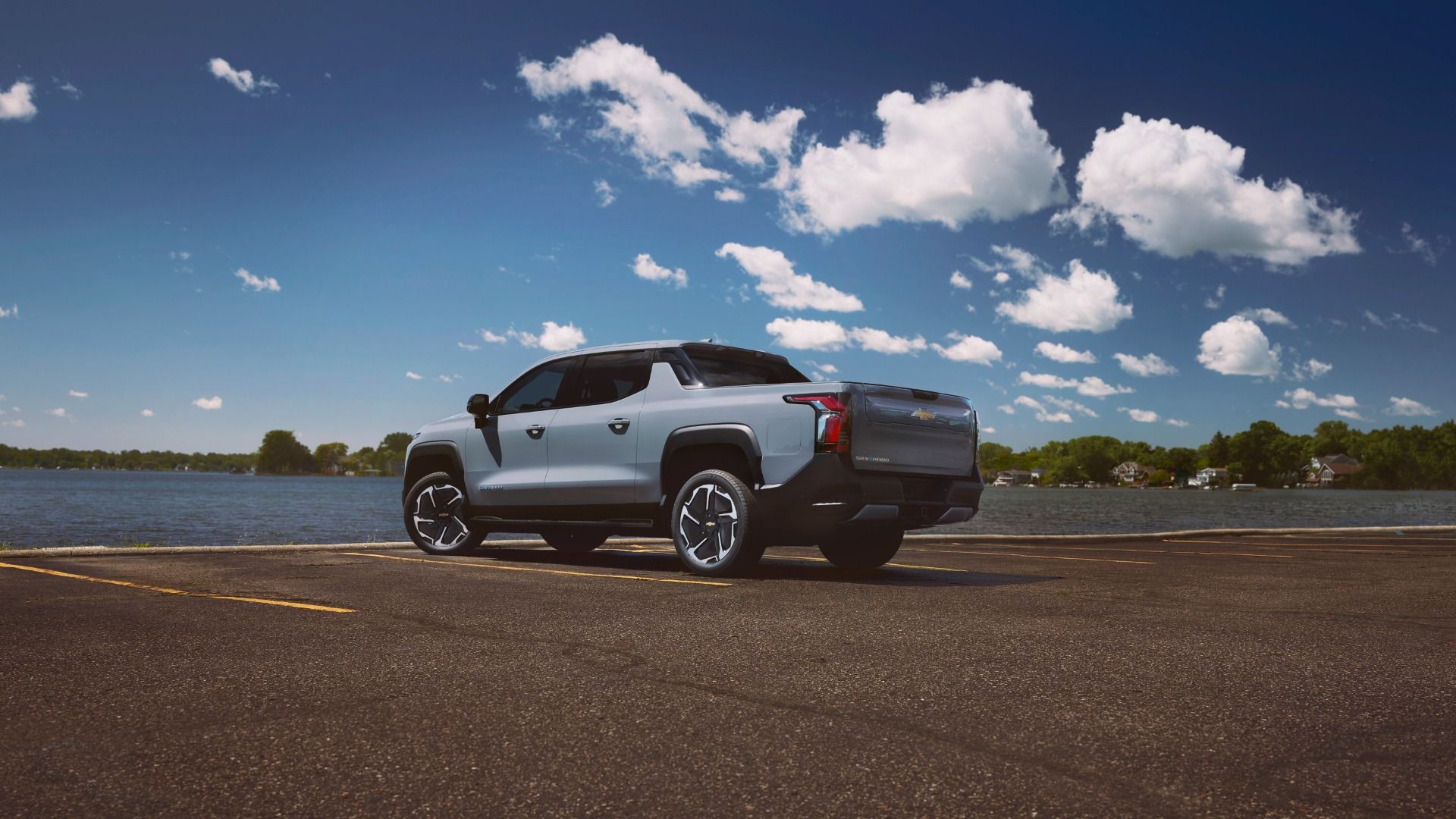 Image of a gray Chevrolet Silverado EV pickup, parked near water, with blue sky and white puffy clouds overhead. 