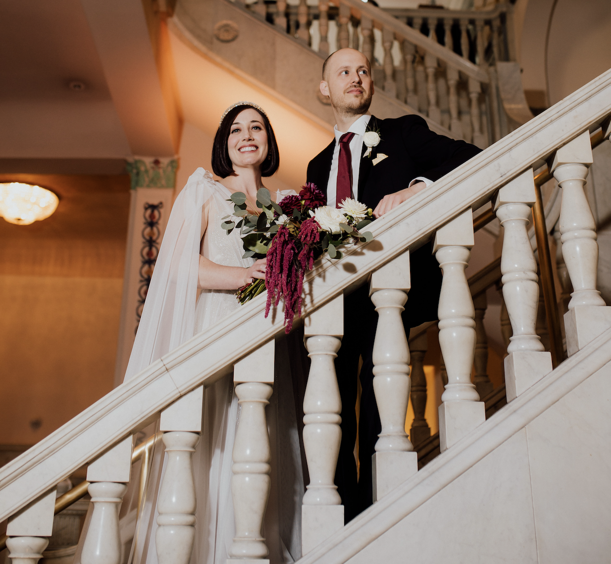 Newlyweds stand on a set of grand stairs and pose for a picture looking over the side.