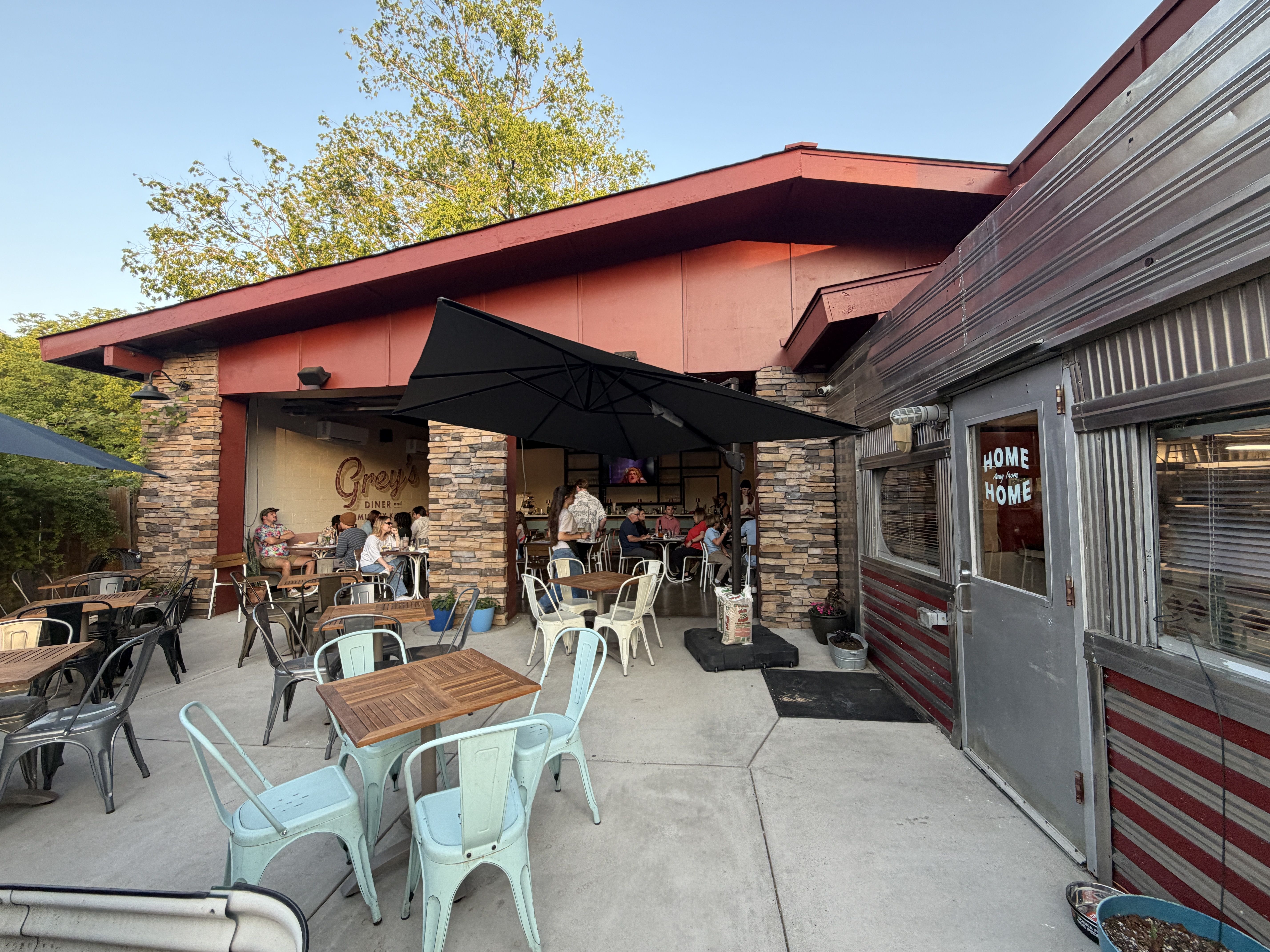 Outdoor diner patio under a red sloped roof with light-blue metal chairs and wooden tables; people dine near stone columns, under a black umbrella, beside a corrugated metal wall.