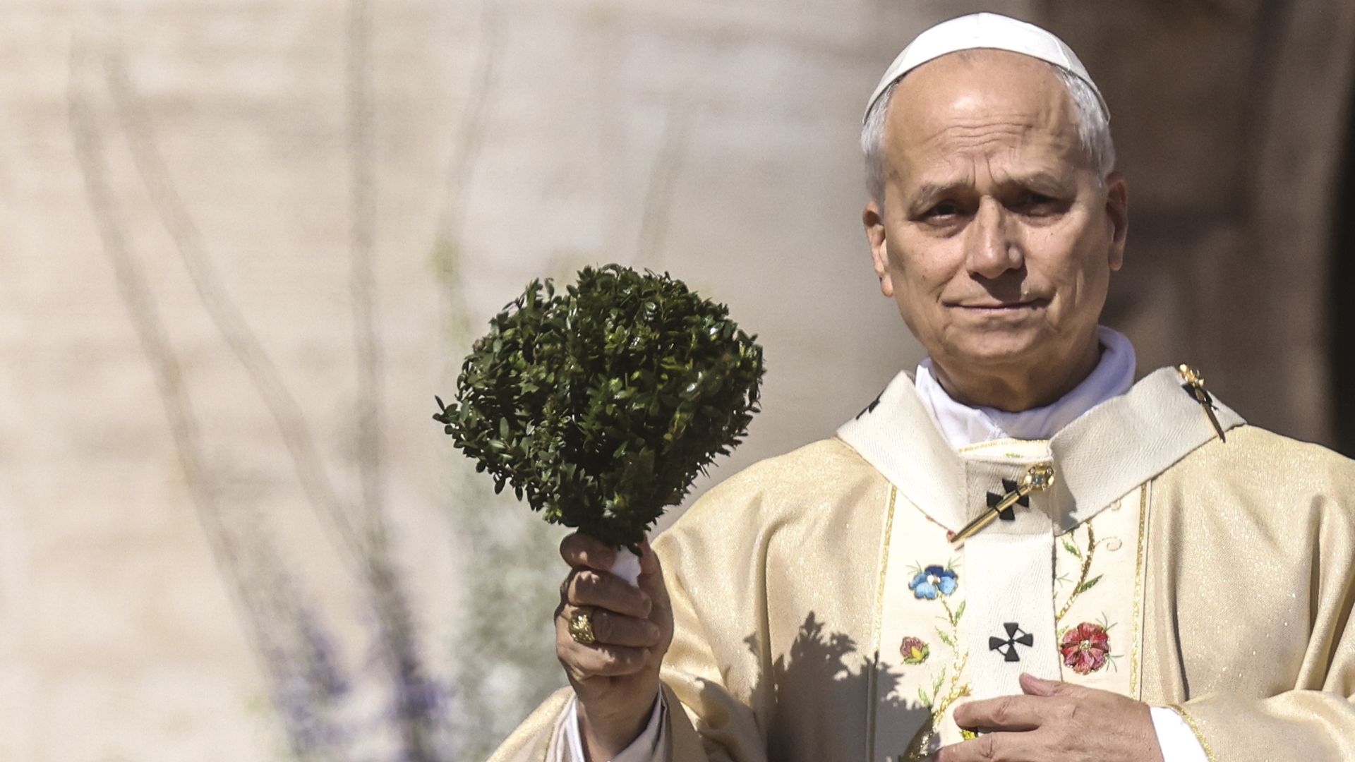 An elderly clergyman in cream liturgical robes with floral embroidery and a white zucchetto, holding a small round green shrub bouquet outdoors against a stone wall.