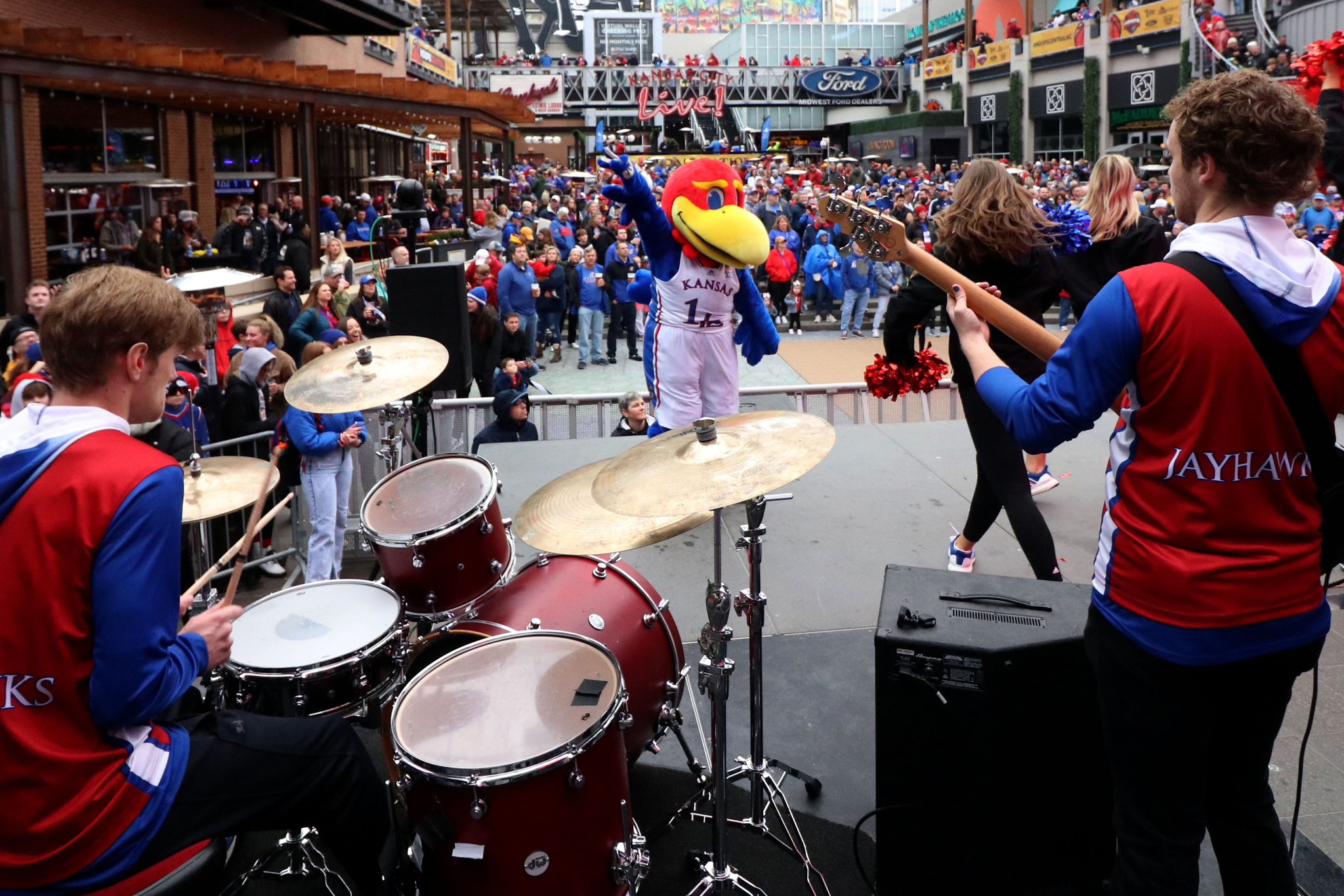 Band performing at outdoor event with Kansas Jayhawks mascot in blue, red, and yellow costume engaging a large crowd wearing blue and red at Kansas City Live venue.