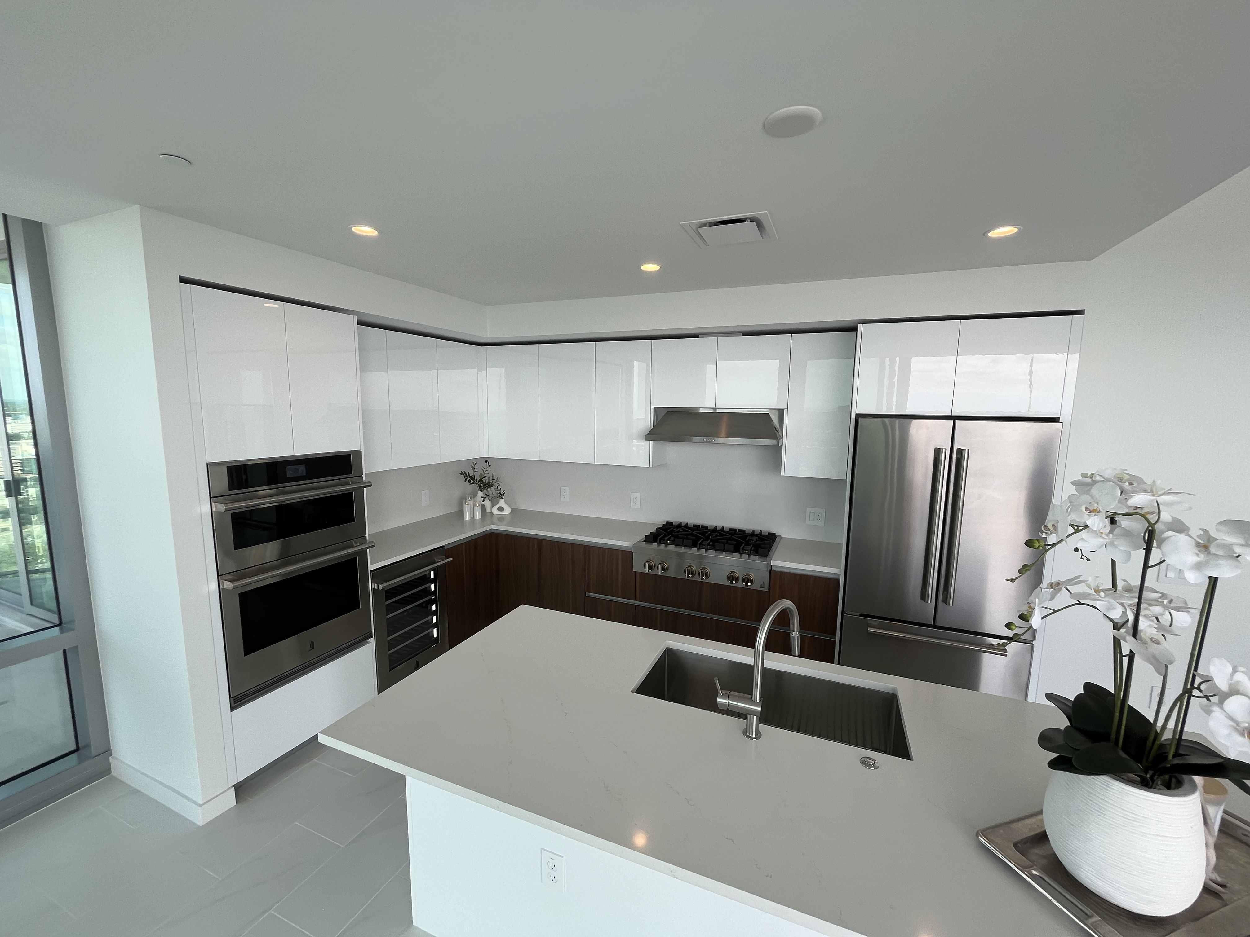 Modern kitchen with white upper cabinets, wooden lower cabinets, stainless steel appliances, large island with sink, and a white orchid plant in a white pot on countertop under recessed ceiling lights.