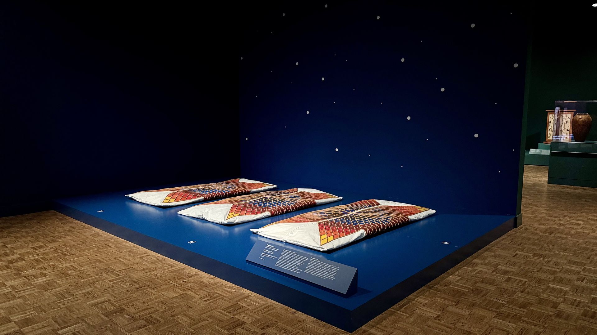 Museum display of sheets of fabric with colorful geometric patterns on a dark blue platform and wall decorated with small white dots resembling stars