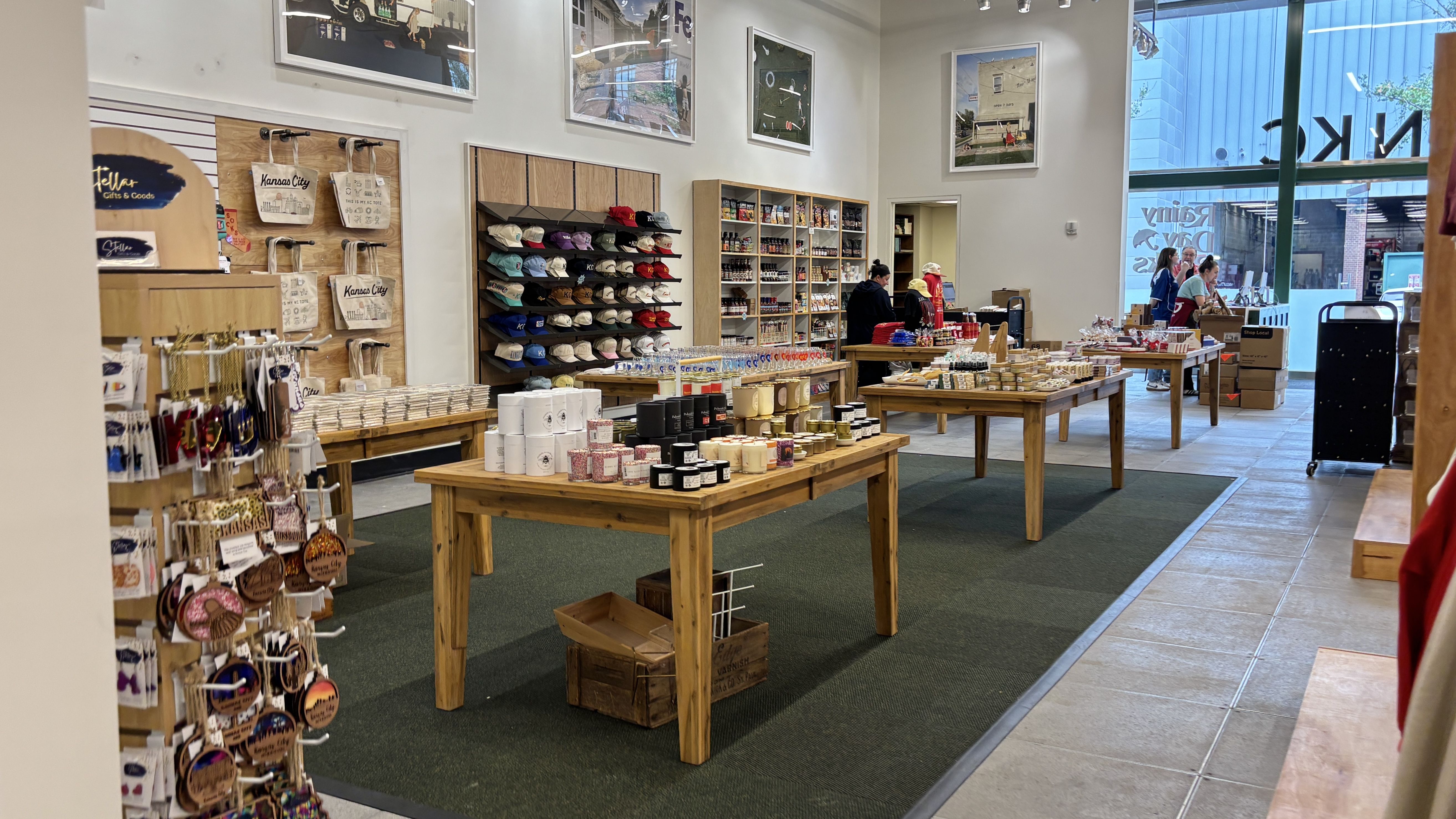 Bright gift shop interior with wooden tables of mugs, candles, and souvenirs. Hat shelves along the back wall under framed prints; customers near the checkout by large windows.