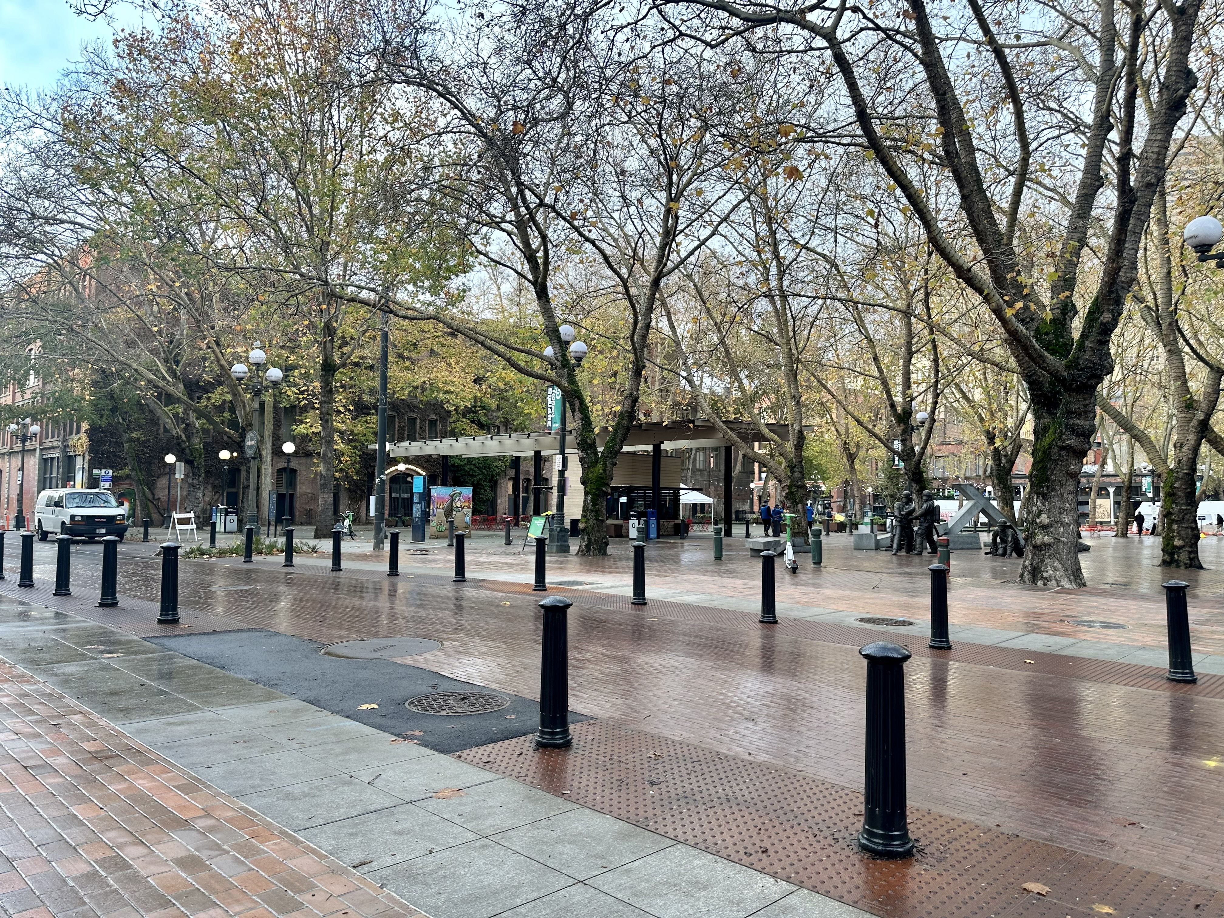 Trees line a paved brick road that opens into a brick plaza, with stanchions bordering the roadway.