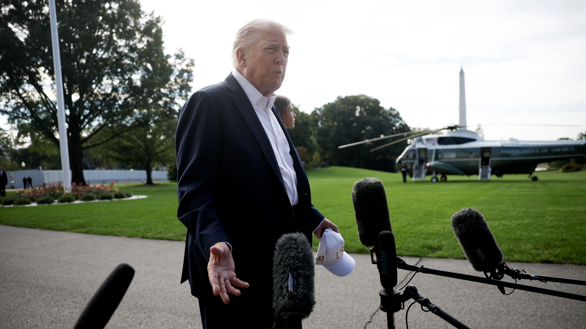 President Trump, wearing a black jacket and white shirt and holding a white baseball cap, speaks to members of the media as he departs the White House. Four large boom mics can be seen in the foreground and Air Force One on the lawn behind him.
