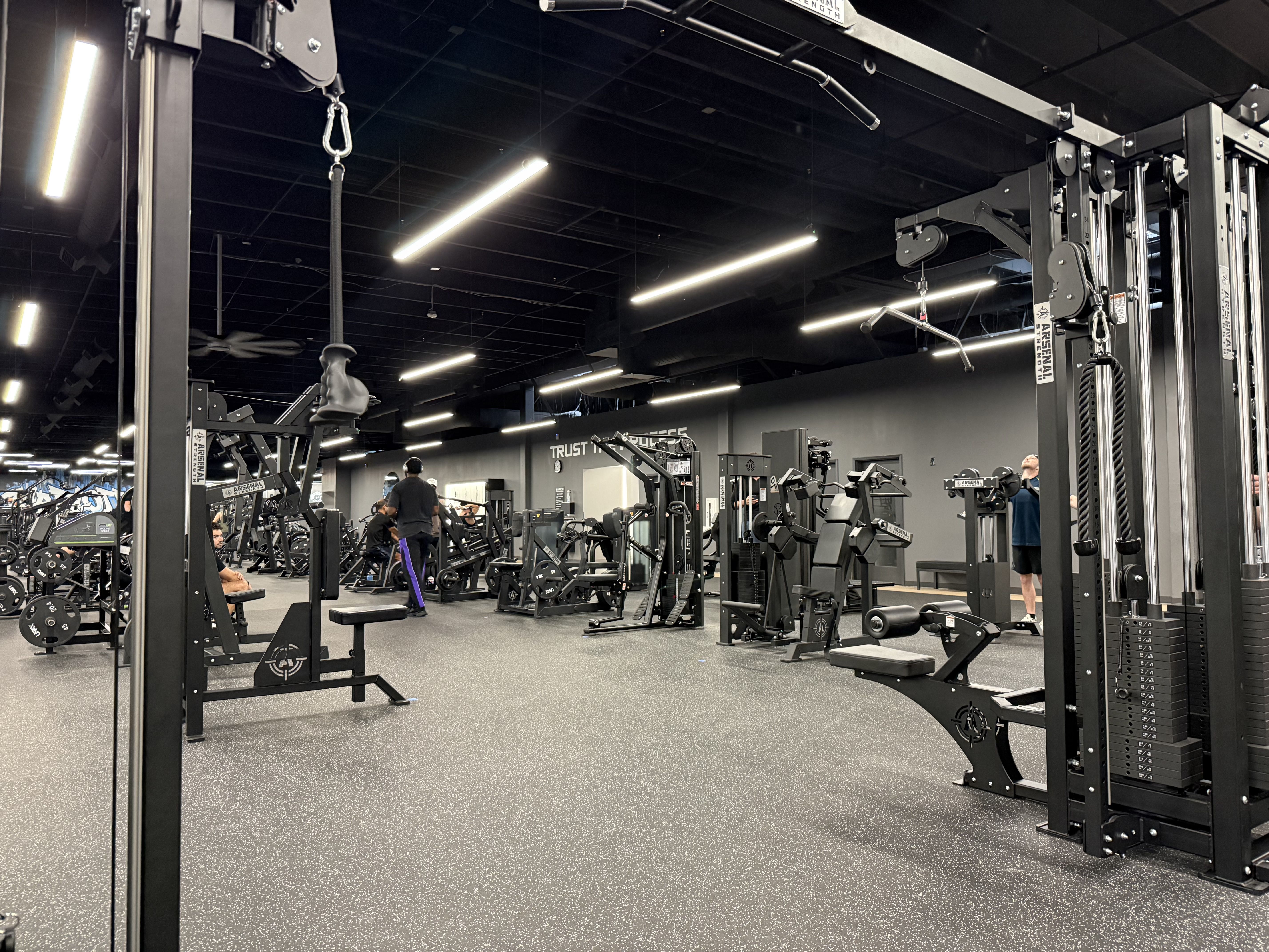 Gym interior with a black ceiling and bright linear lights. A large multi-station weight machine on the right; benches and other equipment fill the space as people work out.
