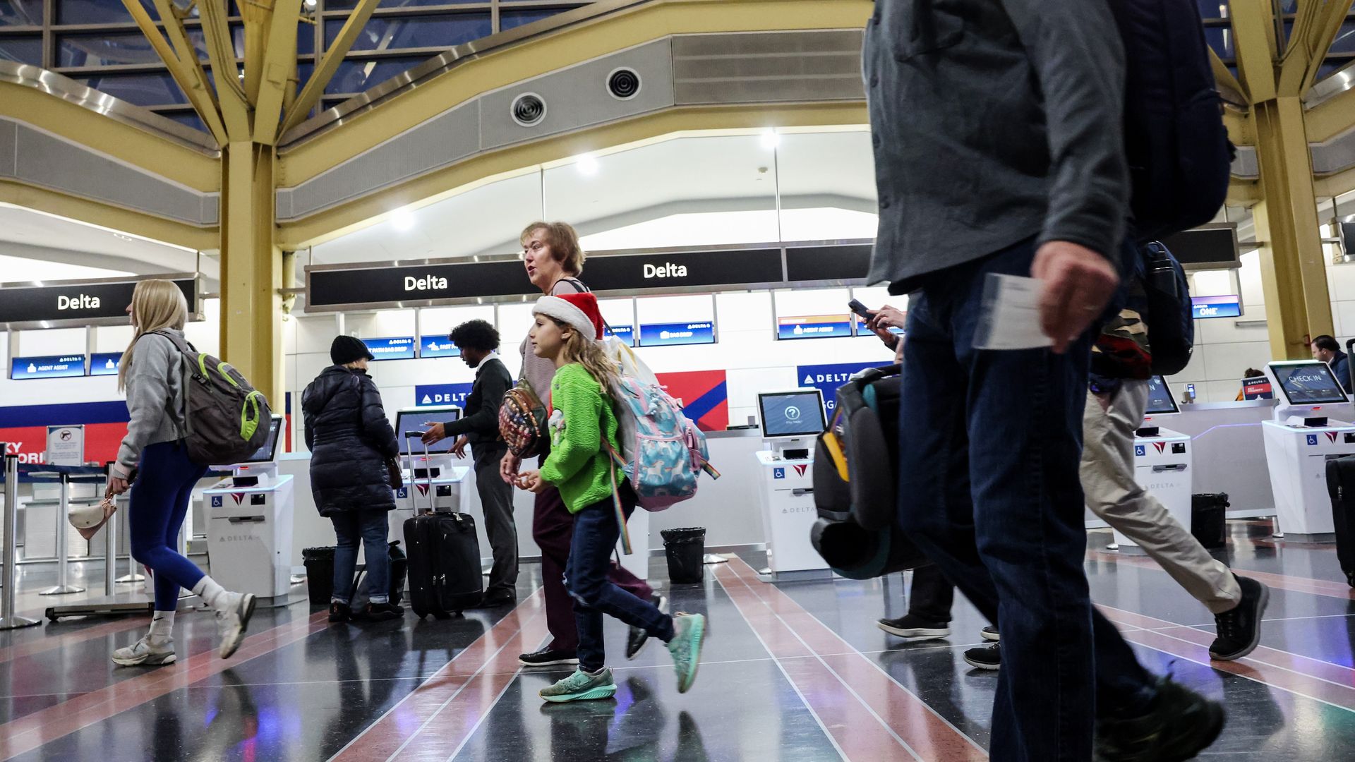 People walking inside an airport terminal near Delta Air Lines check-in counters, with some carrying backpacks and luggage. A child wears a green sweater and a red Santa hat.