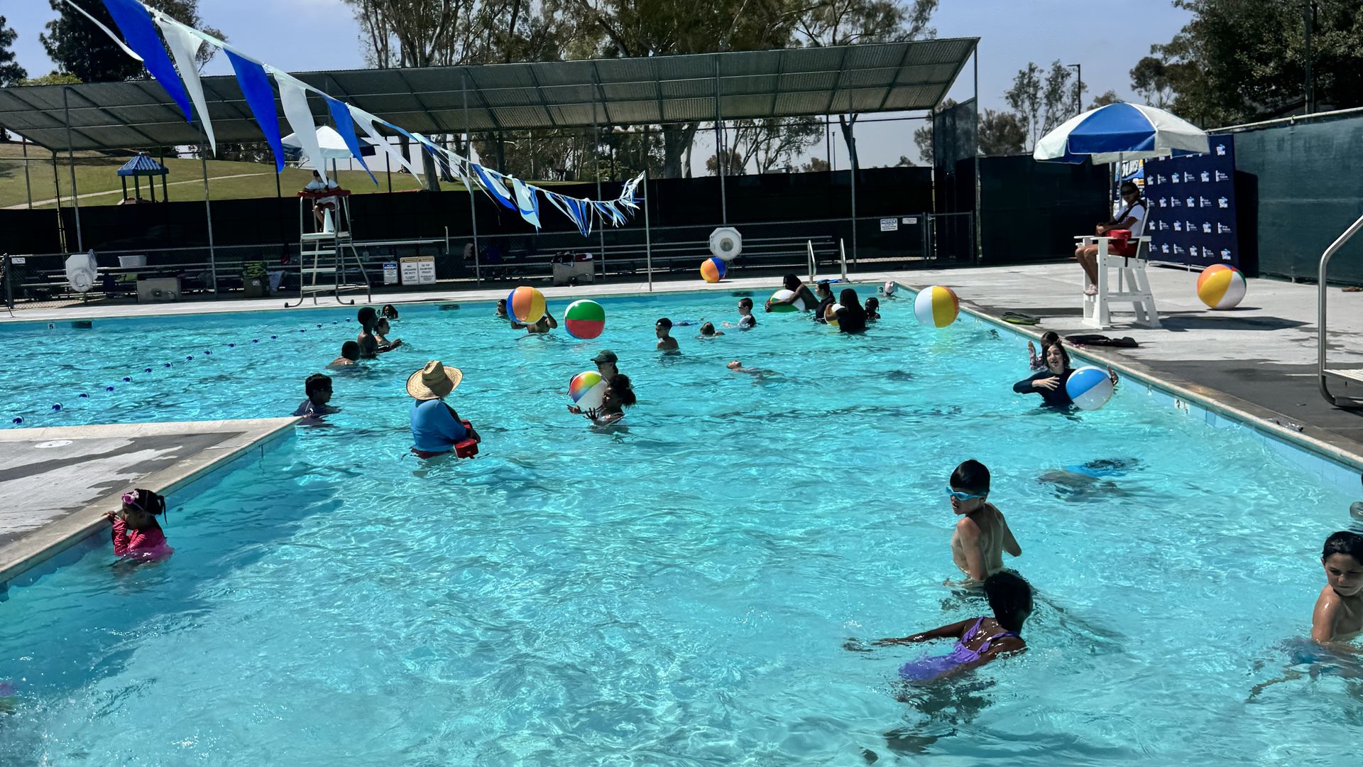 Outdoor swimming pool with kids playing in bright blue water, multicolored beach balls, blue-and-white bunting along a fence, shaded viewing area, umbrellas, and flags in the background.