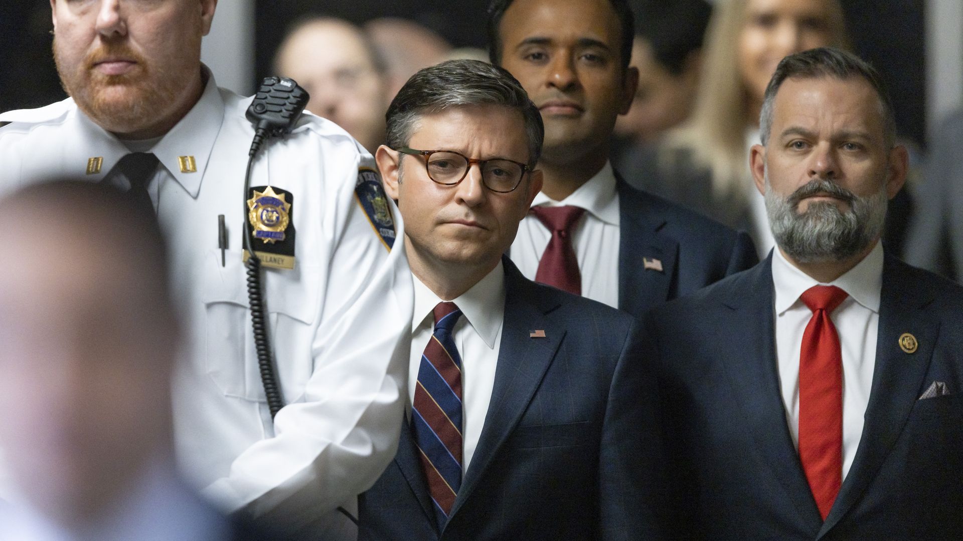 House Speaker Mike Johnson, wearing a blue suit, white shirt, blue-and-white striped tie and glasses, flanked by Rep. Cory Mills, Vivek Ramaswamy and a police officer.