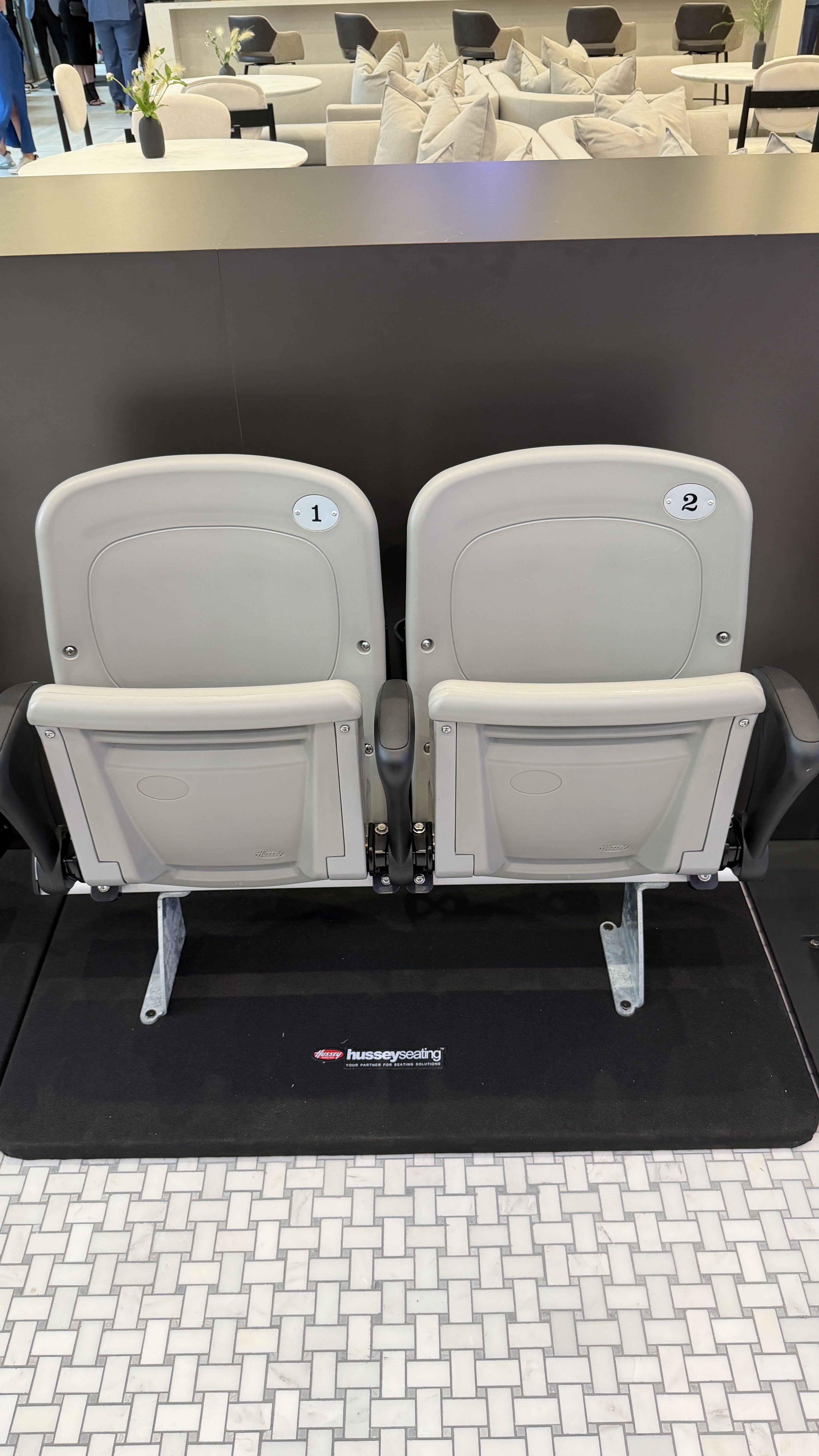 Two light-gray theater seats labeled "1" and "2" on a black platform, facing a modern lounge with white sofas and round tables; a Hussey Seating mat is visible on the floor.