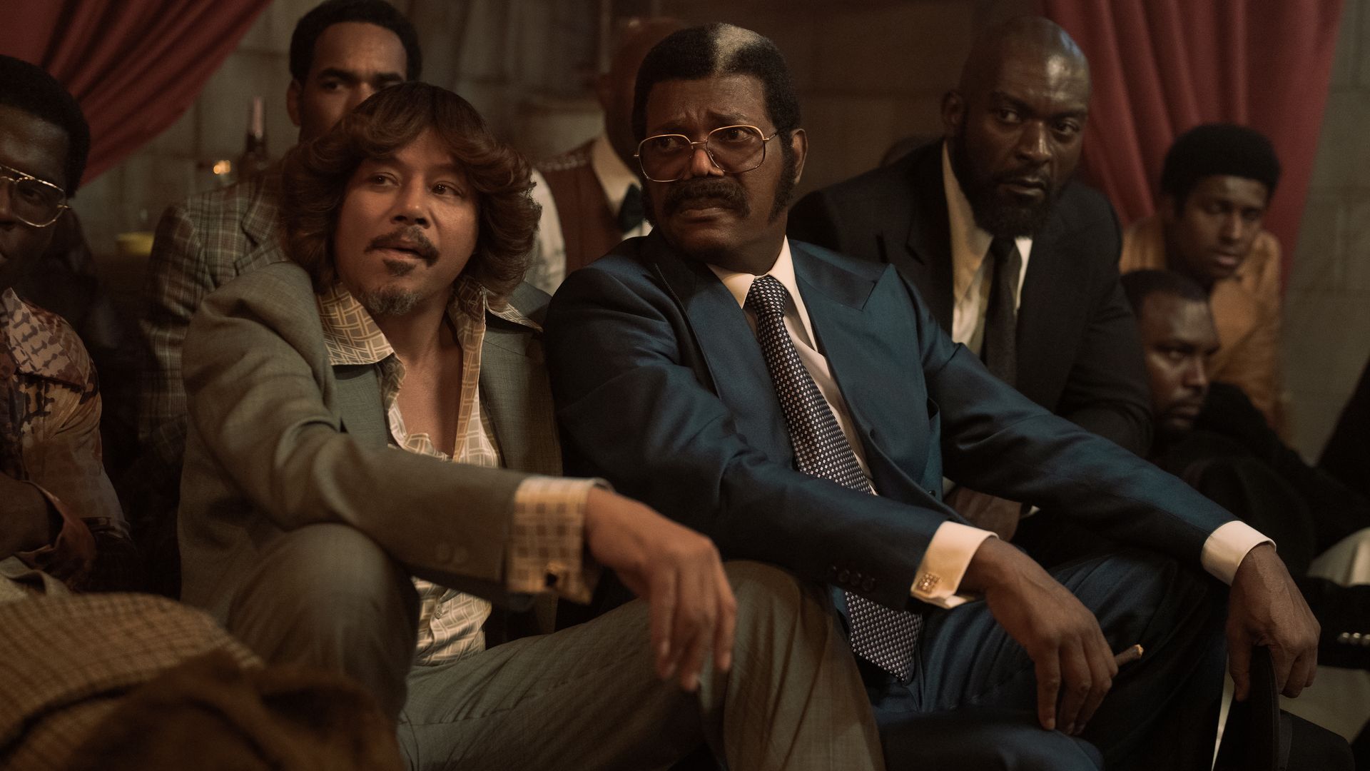 A photo showing a group of men sitting together wearing suits with dated fashions ('70s-style). One has long hair that's flipped out. Another has tinted glasses and a long mustache. 