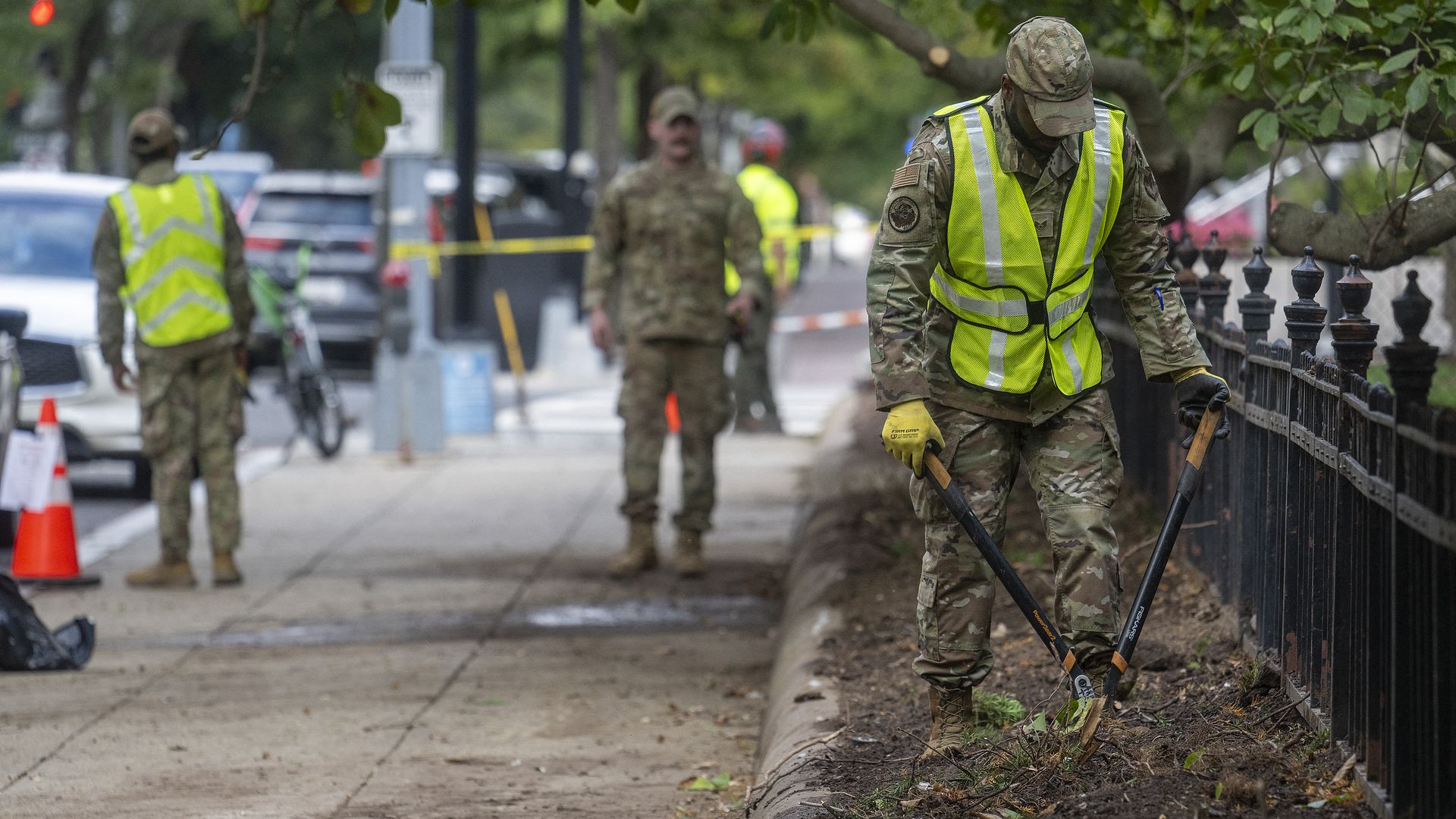 Military personnel in camouflage and neon yellow safety vests work on a sidewalk, one using large pruning shears near a black fence with trees overhead, traffic cones in background.
