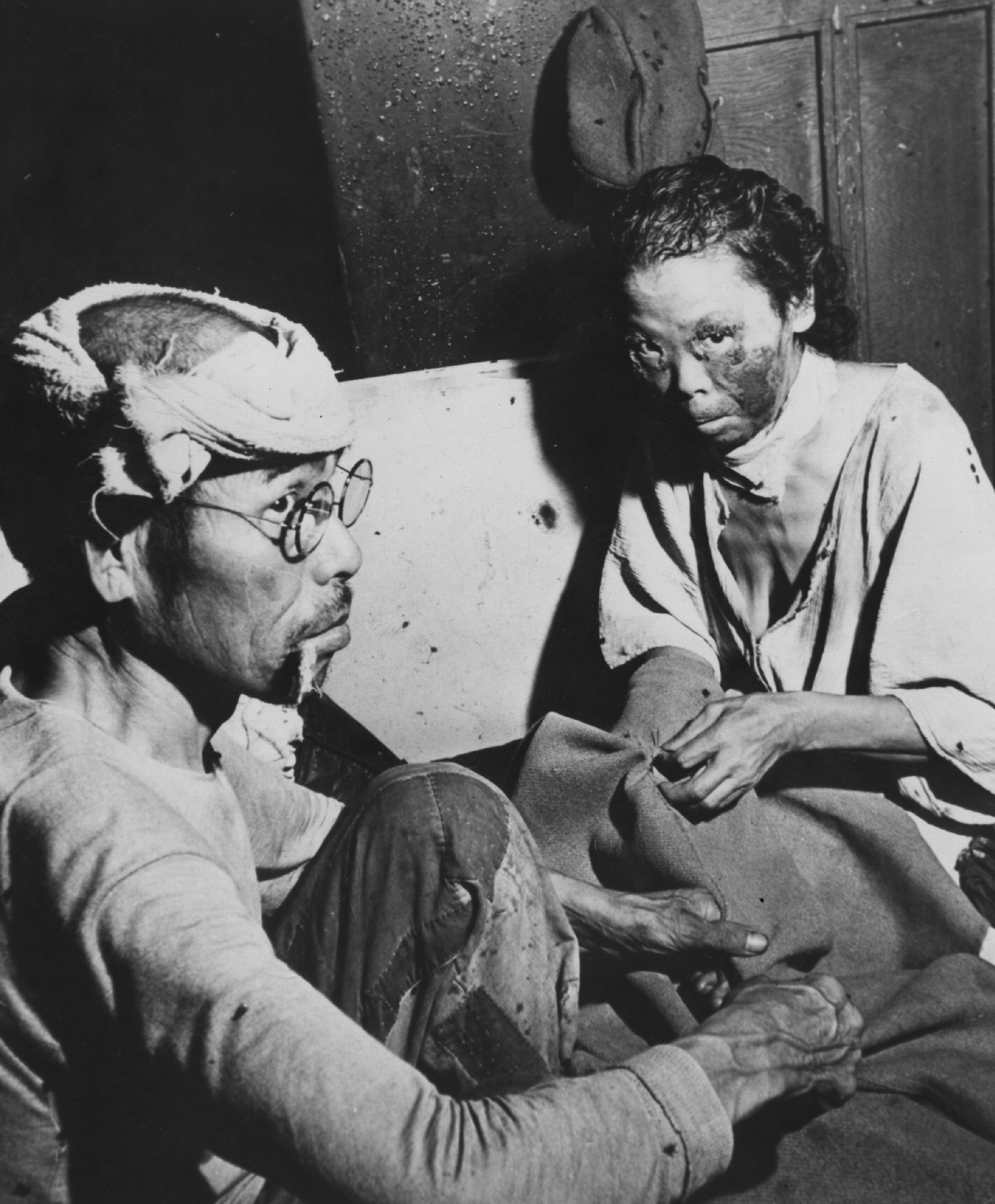 Black and white photo of two Asian individuals sitting closely; one wears glasses and a head wrap while the other has facial scars and is holding fabric, against a wooden wall background.