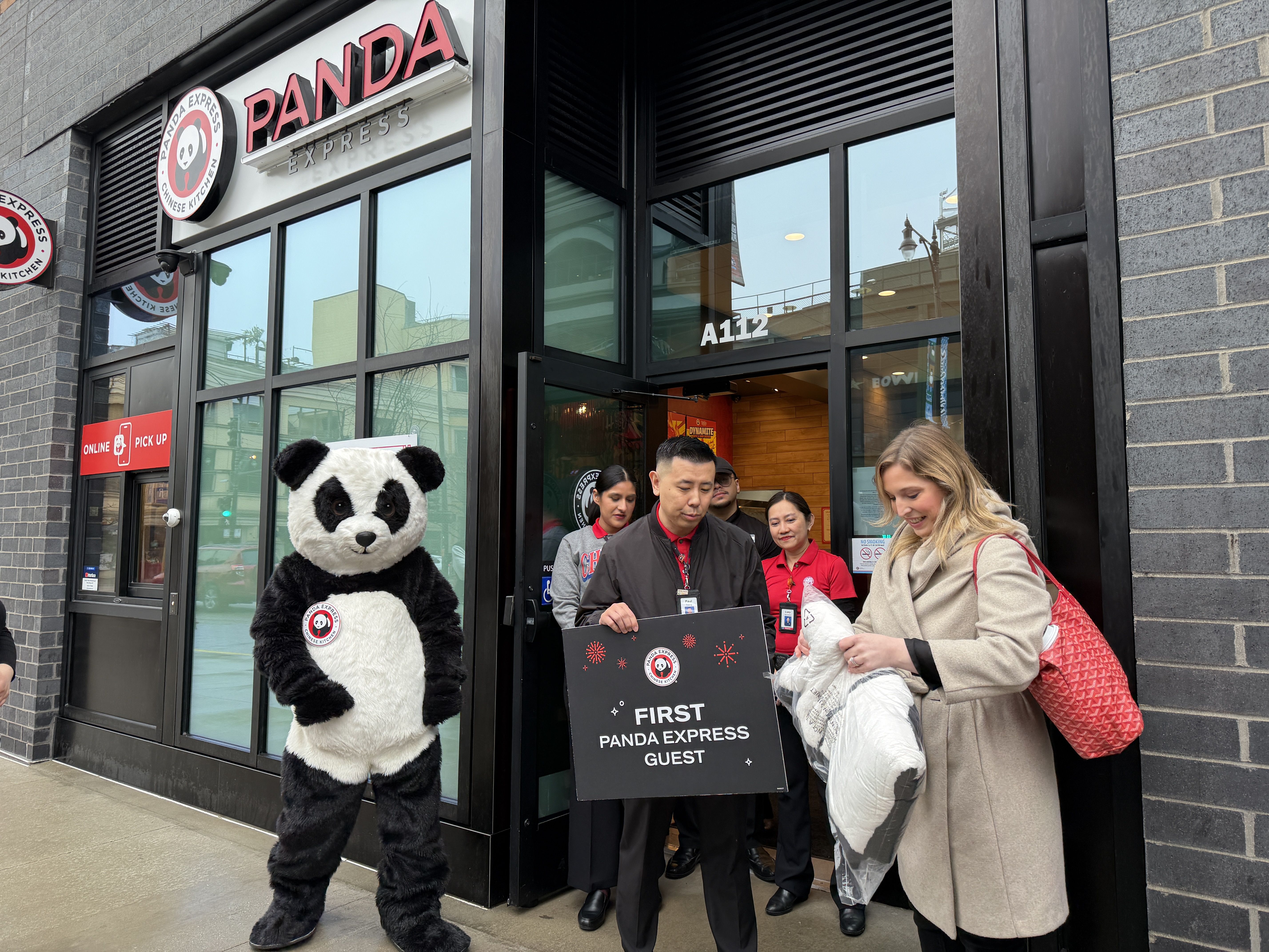 People stand outside a Panda Express restaurant, one in a panda costume, a man holding a sign reading "FIRST PANDA EXPRESS GUEST," and a woman smiling with a red handbag.
