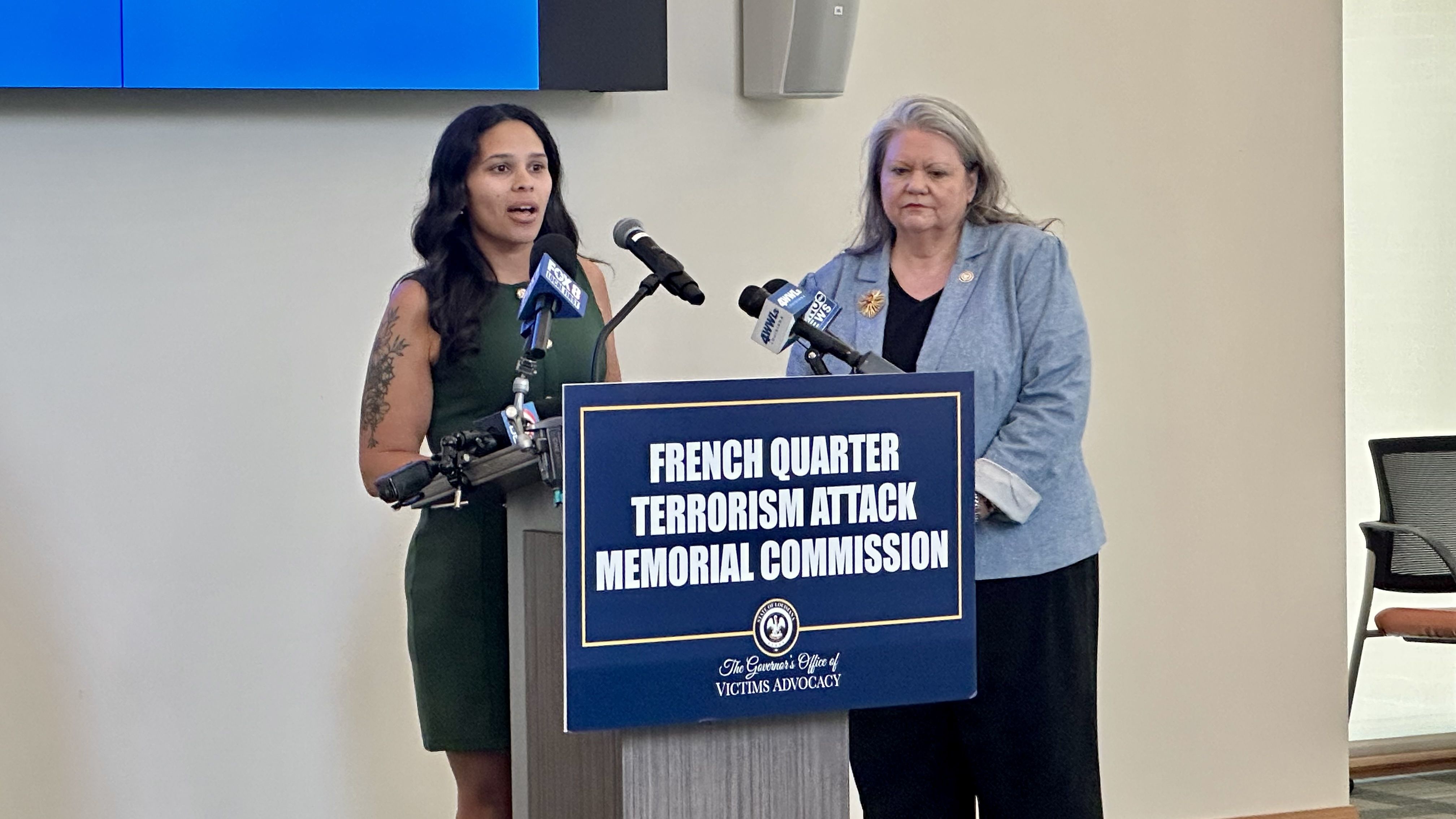Two women speak at a press conference behind a podium with microphones; the lectern reads "FRENCH QUARTER TERRORISM ATTACK MEMORIAL COMMISSION." One wears a green dress with a tattoo on her arm, the other a light blue blazer.