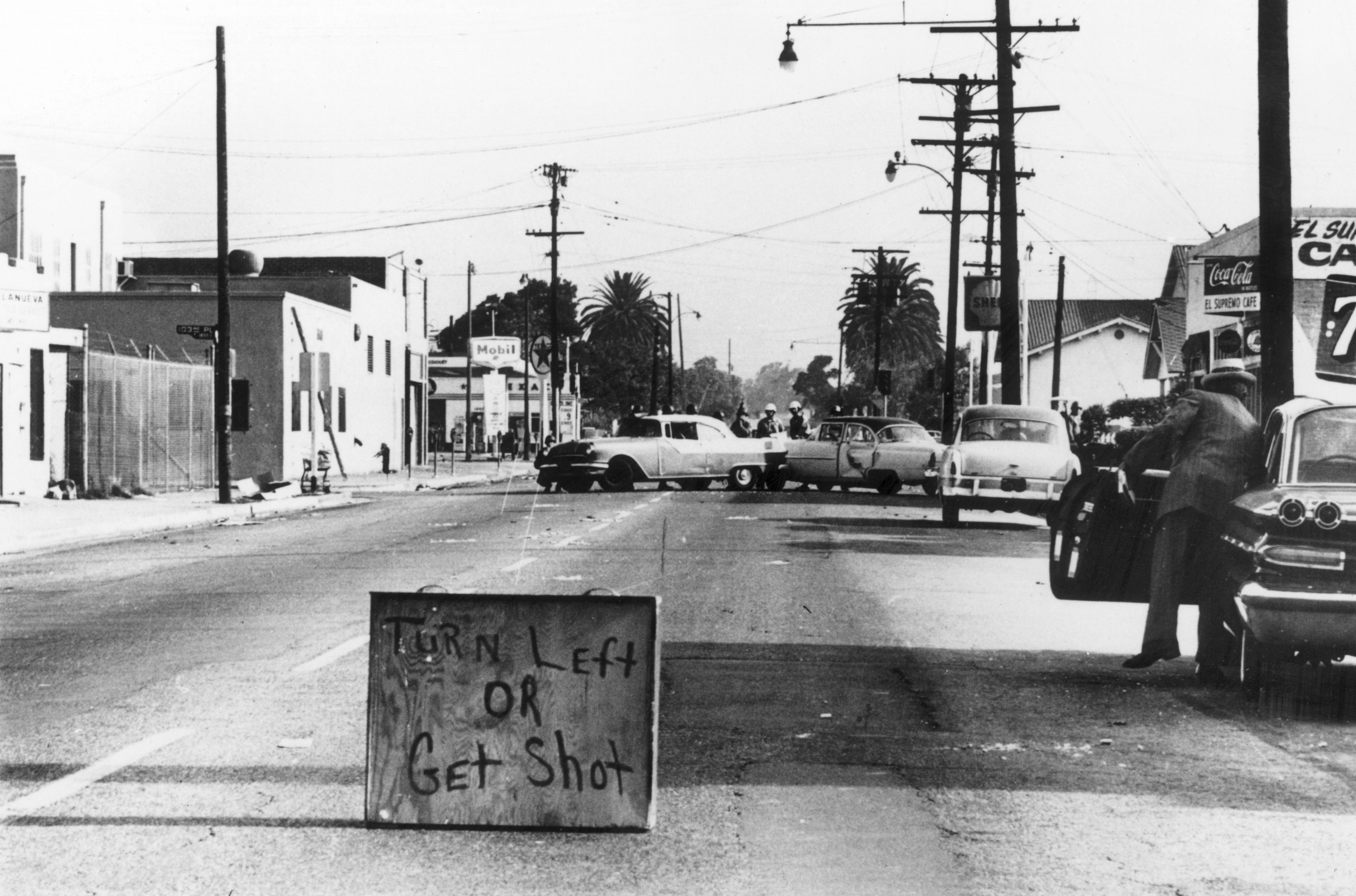 Black and white photo of a street with a wooden sign saying "TURN LEFT OR GET SHOT." Several vintage cars block the road, with palm trees and utility poles in the background.