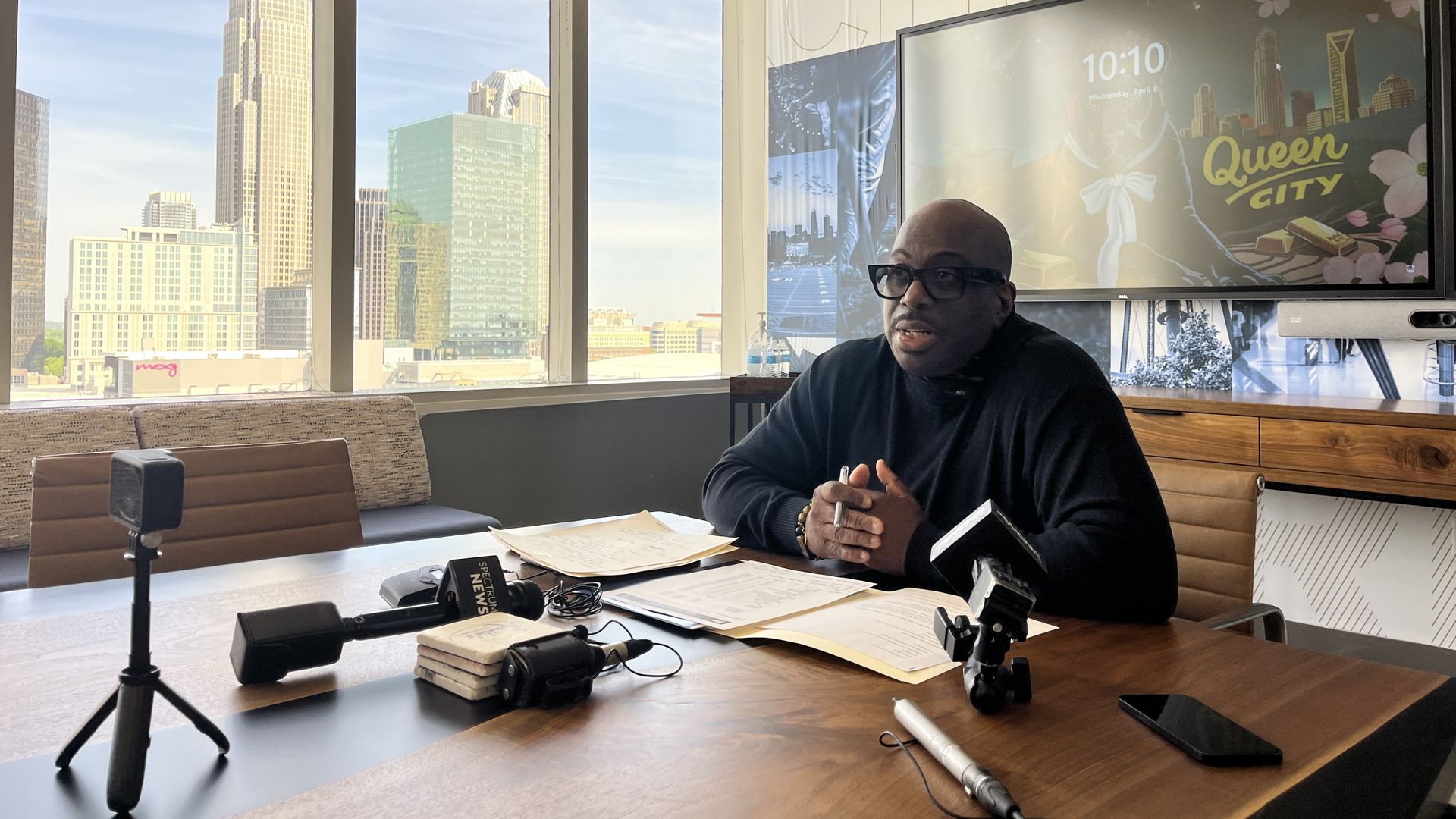 Bald man in a black turtleneck and glasses sits at a wooden conference table in a bright office. He clasps his hands; microphones and papers lie nearby as a city skyline is visible through large windows, and a screen reads "Queen City".